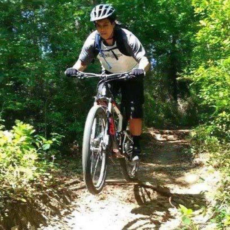 A cyclist wearing a helmet and sporty attire performs a jump on a mountain bike while riding along a wooded trail. Lush greenery surrounds the path, creating a vibrant outdoor environment. UWF Mountain Bike Trails mountain bike trail.