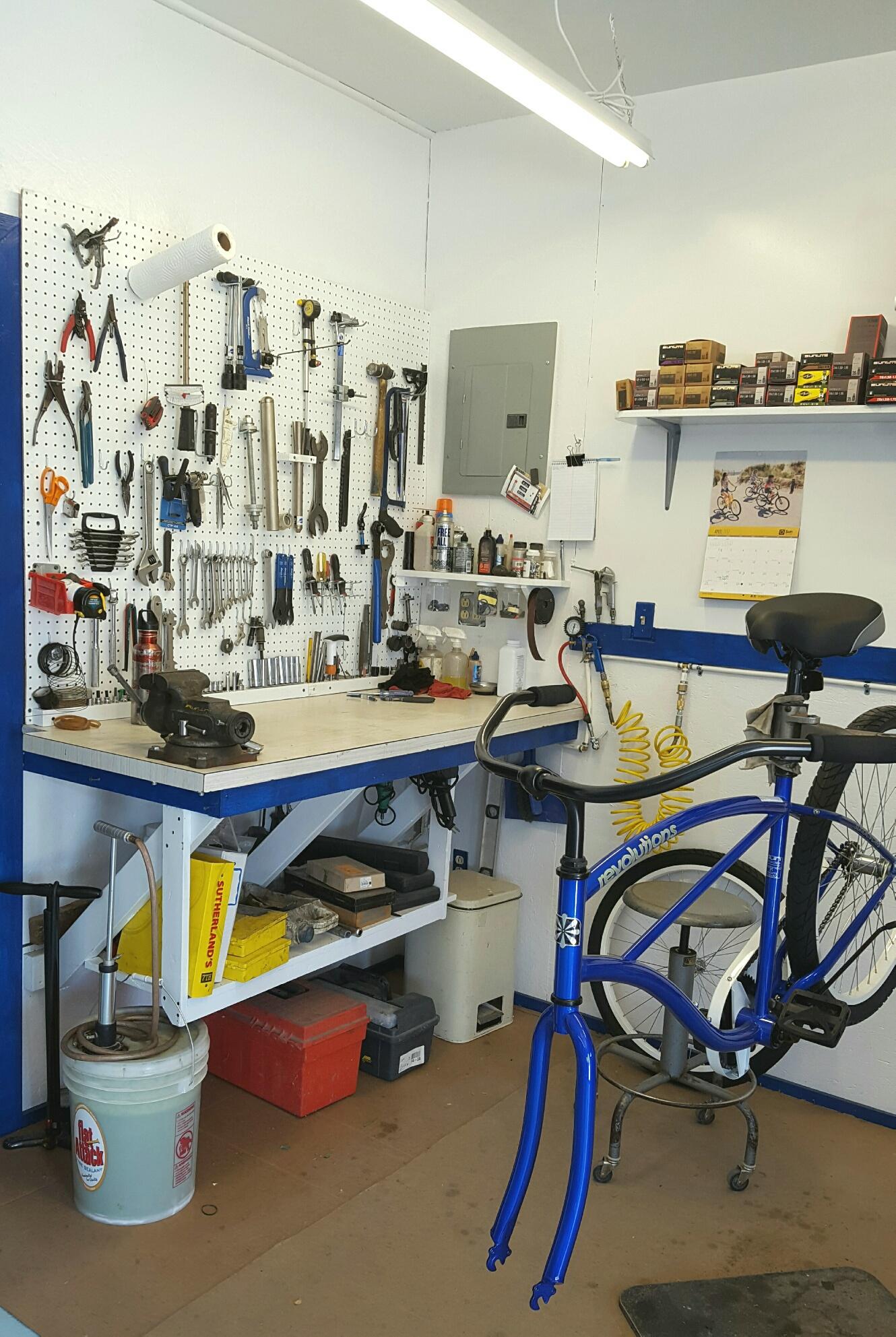 A bicycle repair workspace featuring a well-organized pegboard with various tools including wrenches, pliers, and a drill. A wooden workbench is in the foreground, with a bicycle frame partially assembled and tools scattered around. Shelves above hold additional bike parts and supplies, with a calendar hanging on the wall. The room has a bright overhead light and a clean, functional atmosphere.