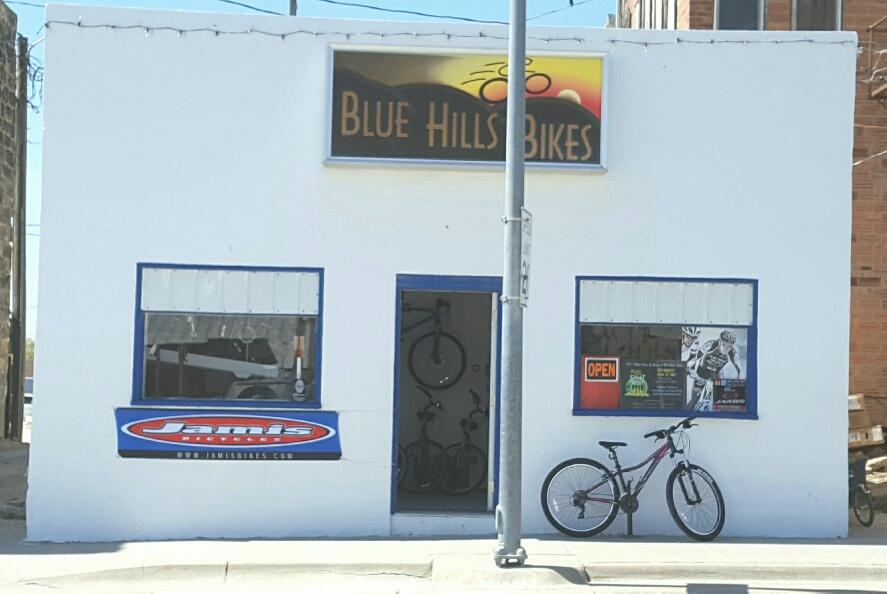 A small white building with a sign reading "Blue Hills Bikes" at the top. The front features a doorway with bicycles visible inside and an "OPEN" sign in the window. A blue banner displaying "Jamis Bicycles" is below one window, and a bicycle is parked beside the building.