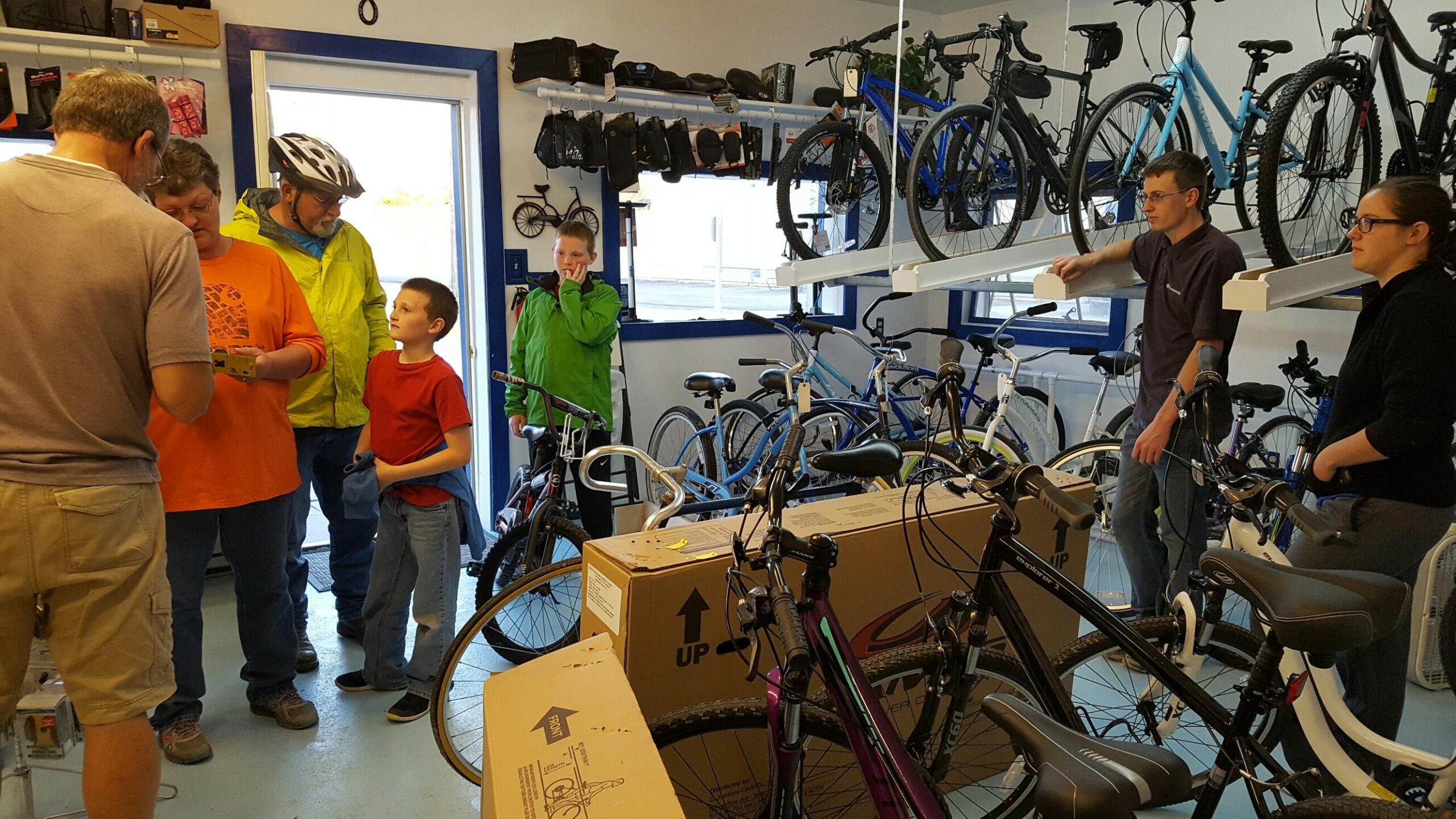 A group of people, including adults and children, gathered inside a bike shop surrounded by various bicycles and accessories. One adult is wearing a helmet, while another is handling items with two young boys nearby. Shelves hold additional bikes and gear, and a boxed bike is visible in the foreground. The atmosphere appears busy and engaging as customers interact with staff.