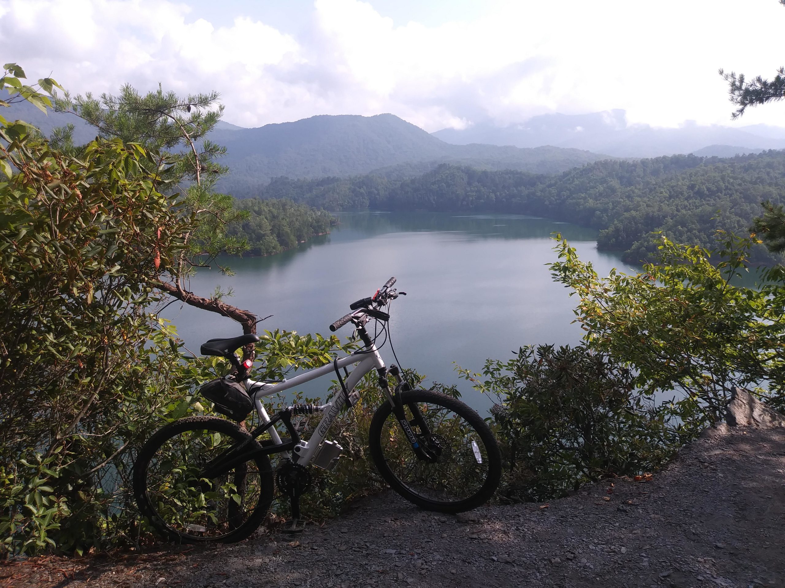 A mountain bike resting on a gravel path beside a serene lake, surrounded by lush greenery and distant mountains under a partly cloudy sky. Tsali Right Loop mountain bike trail.