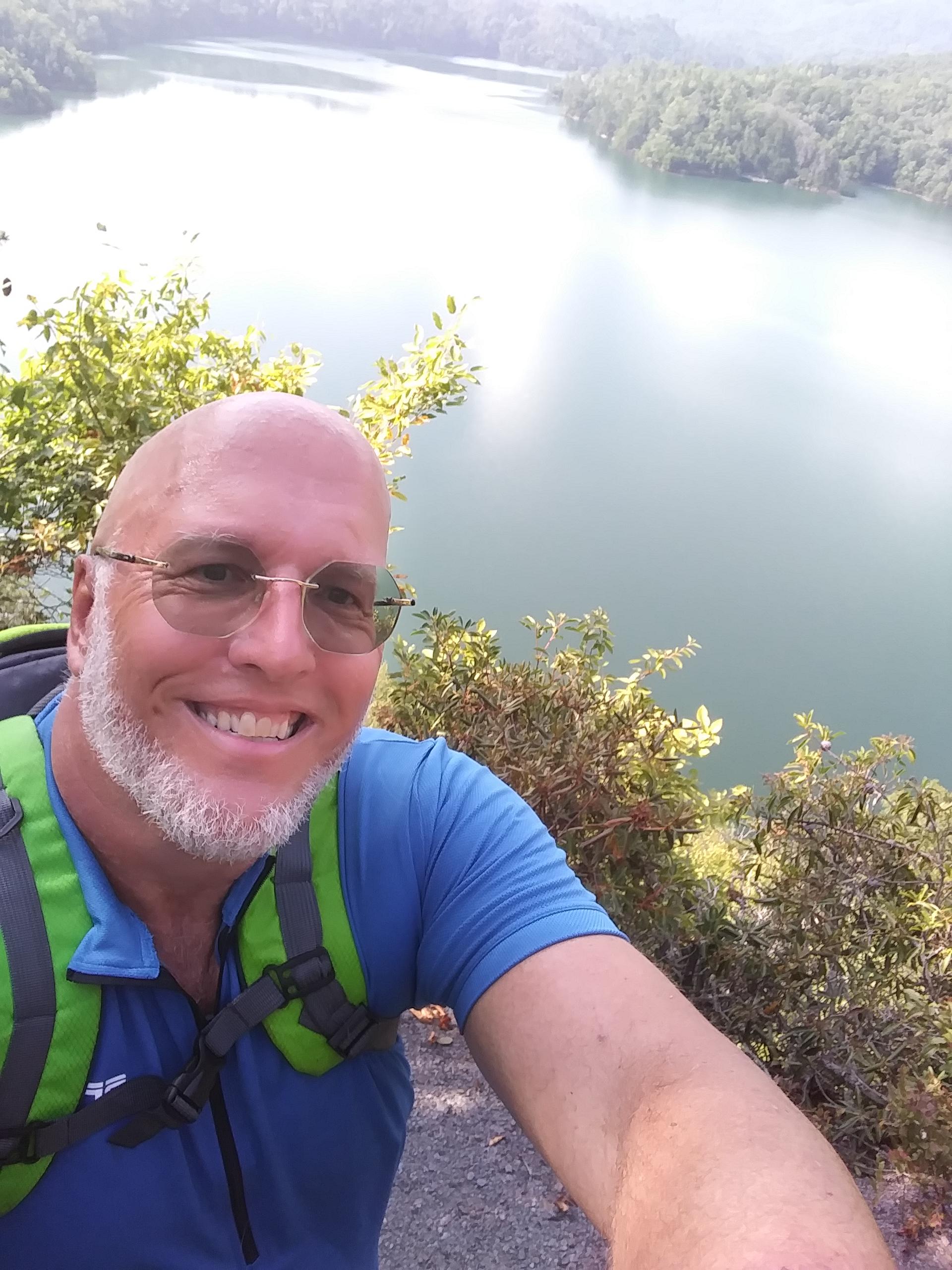 A smiling person with short, bald hair and a beard is posing for a selfie, wearing glasses and a blue shirt with a green backpack. In the background, a serene lake surrounded by trees and mountains is visible, reflecting the sunlight and blending with the natural scenery. Tsali Left Loop mountain bike trail.