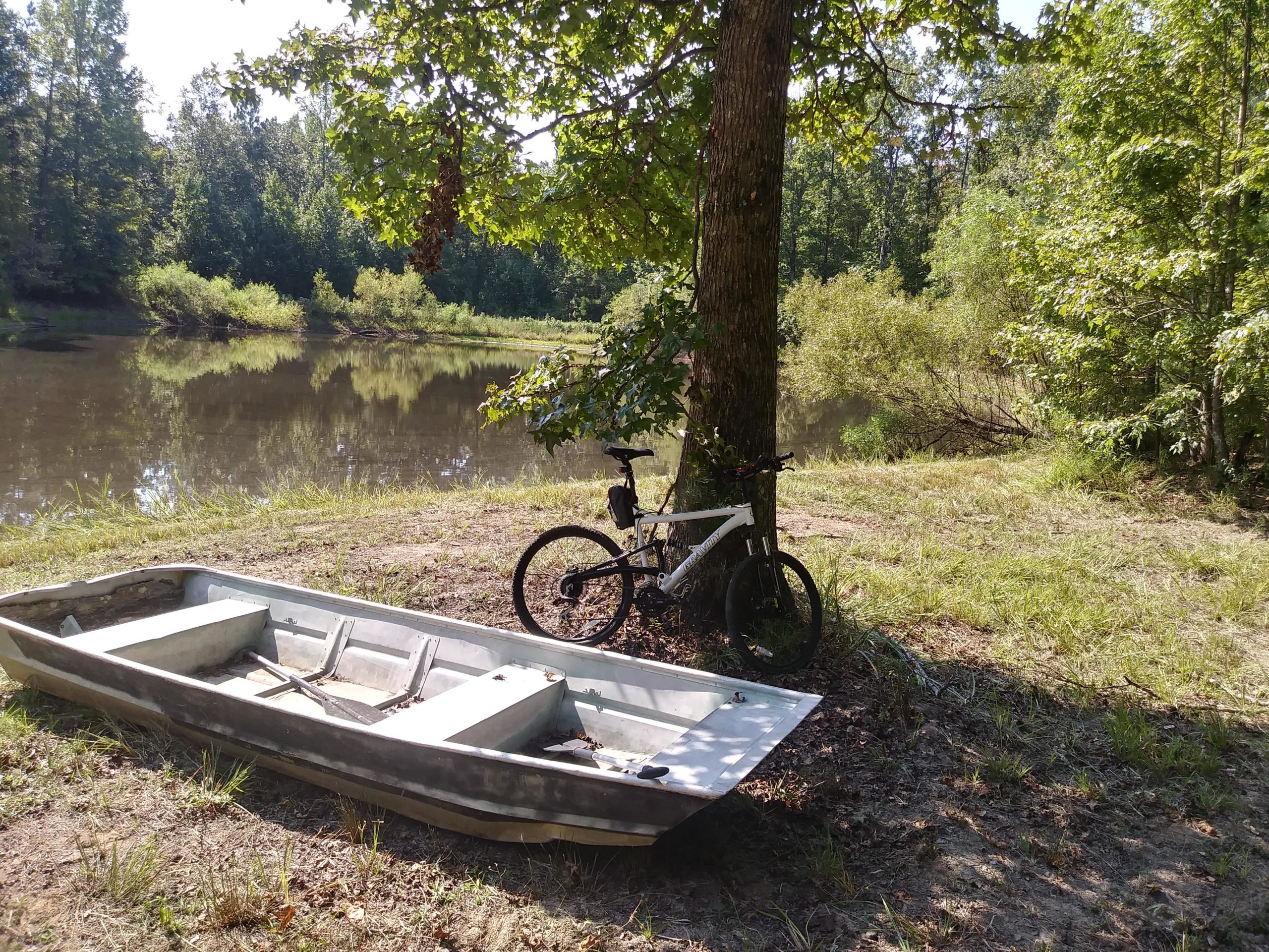 A serene outdoor scene featuring a calm river with reflections of greenery. In the foreground, there is a weathered fishing boat resting on the grass beside the water. Nearby, a mountain bike is leaning against a tree, surrounded by lush foliage and a peaceful natural landscape. Tannehill Historic Ironworks State Park mountain bike trail.