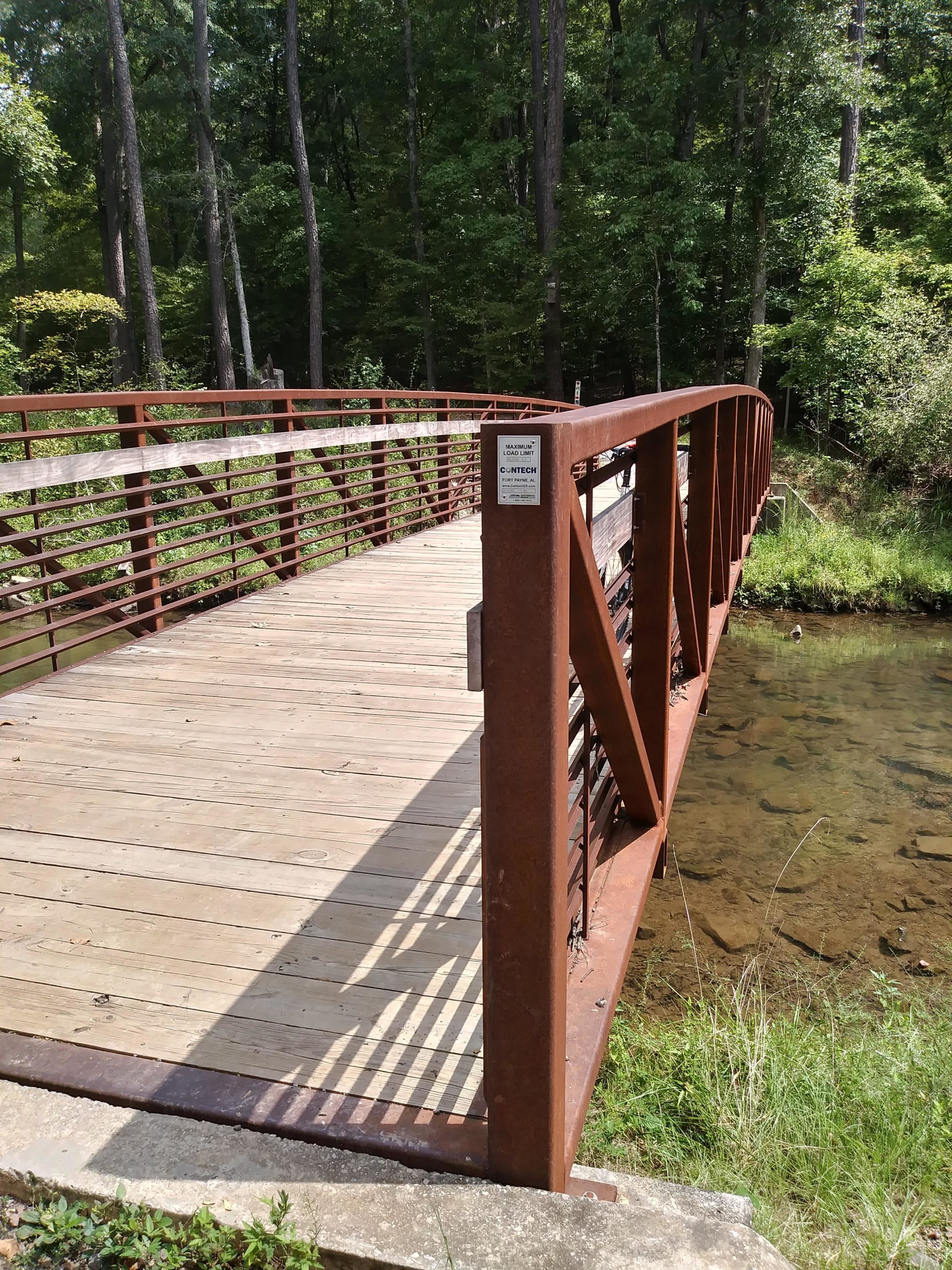 A metal bridge with a wooden deck spanning over a clear stream, surrounded by lush greenery and trees. The bridge features a rust-colored railing and a sign indicating weight capacity. Sunlight casts shadows on the bridge, highlighting its structure. Tannehill Historic Ironworks State Park mountain bike trail.