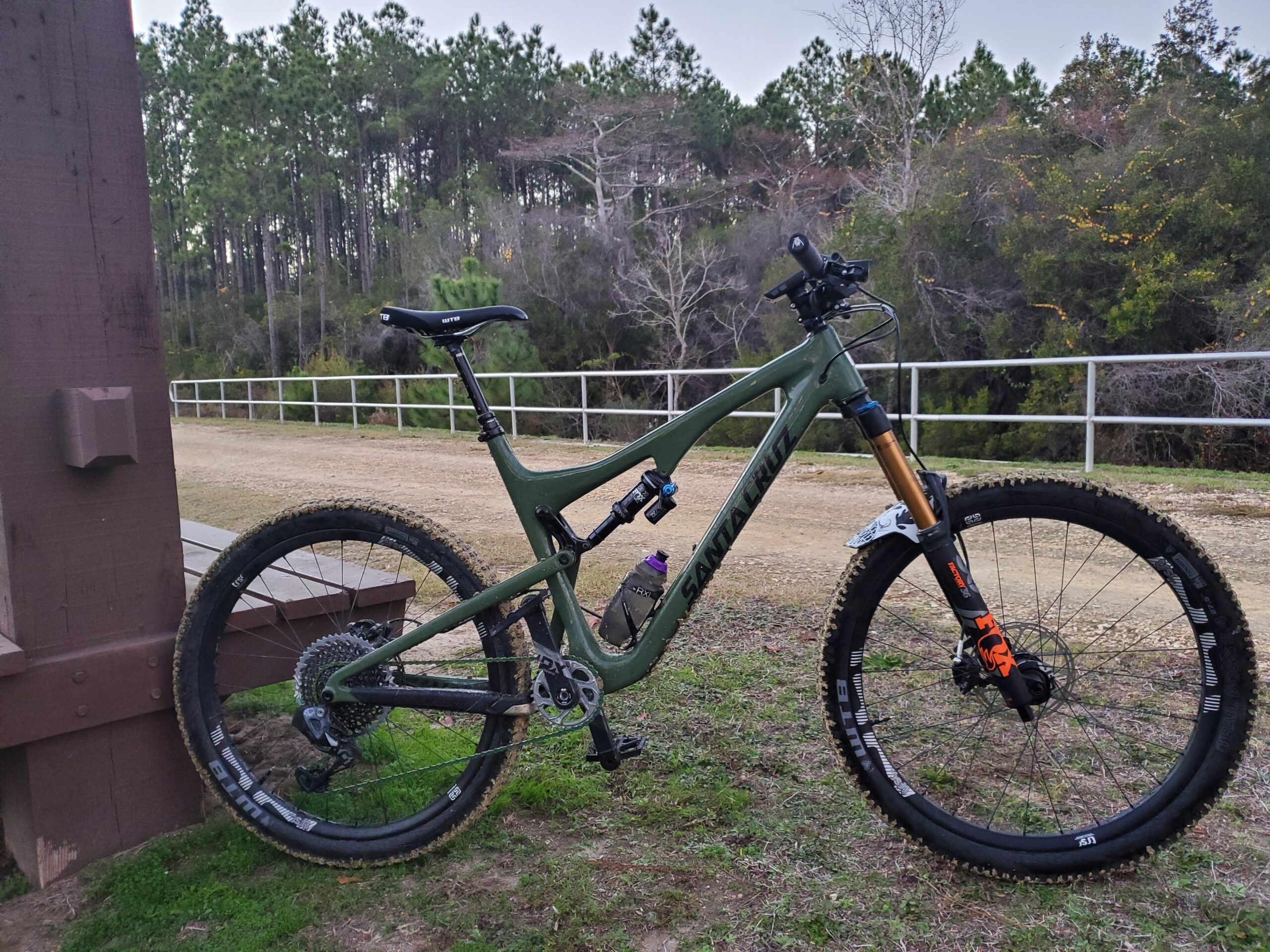 Santa Cruz Bronson: A green mountain bike is parked beside a wooden structure, with its front wheel on a grassy area and the background featuring a dirt path and tall pine trees. The bike has sturdy tires showing some mud, and the scene is set in a natural outdoor environment during twilight.