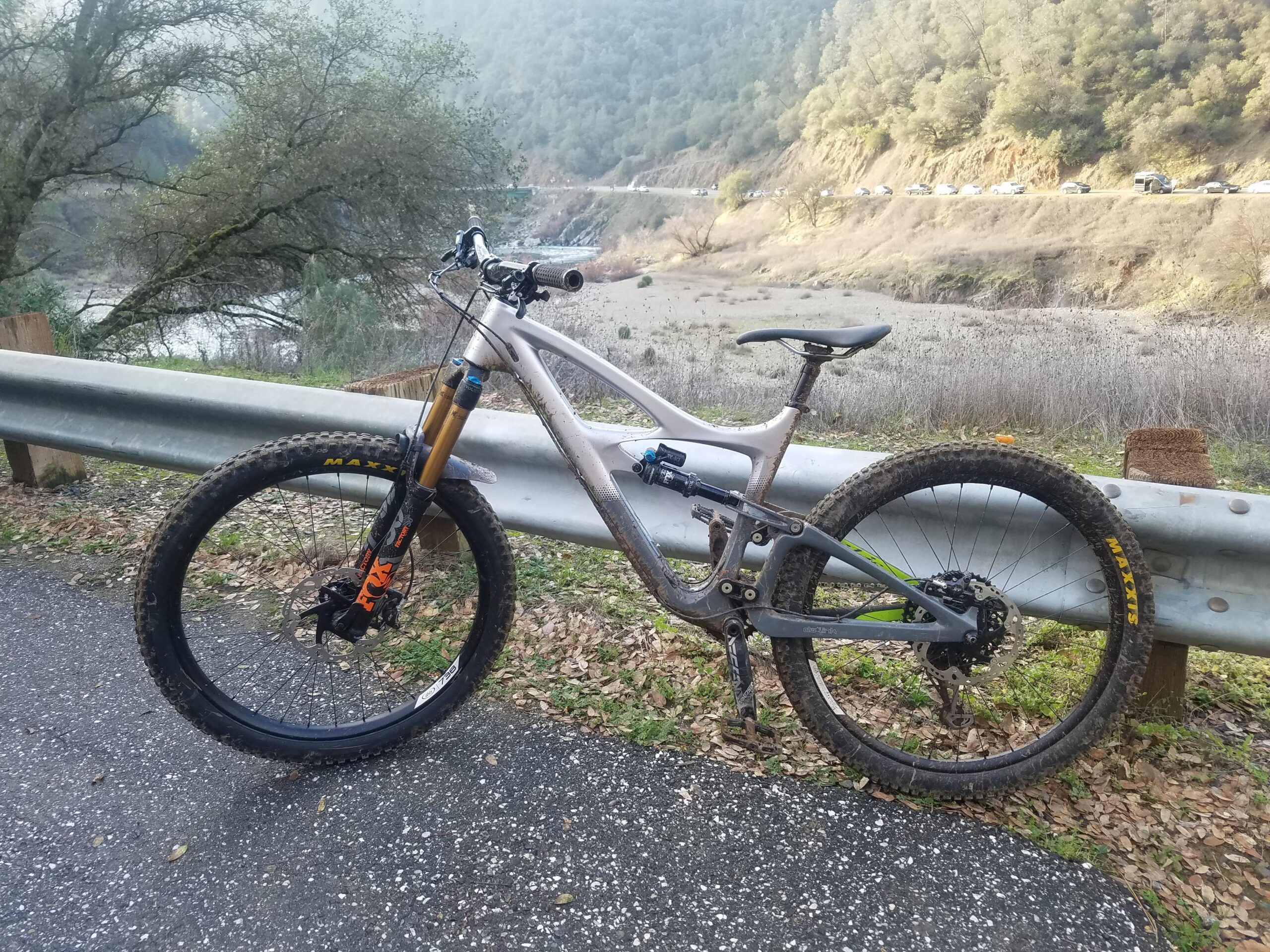Ibis Mojo HD4: A mountain bike leaning against a guardrail on a rural road. In the background, a scenic view of a river and rolling hills can be seen, with trees lining the banks. The road surface is paved with some leaves scattered around.