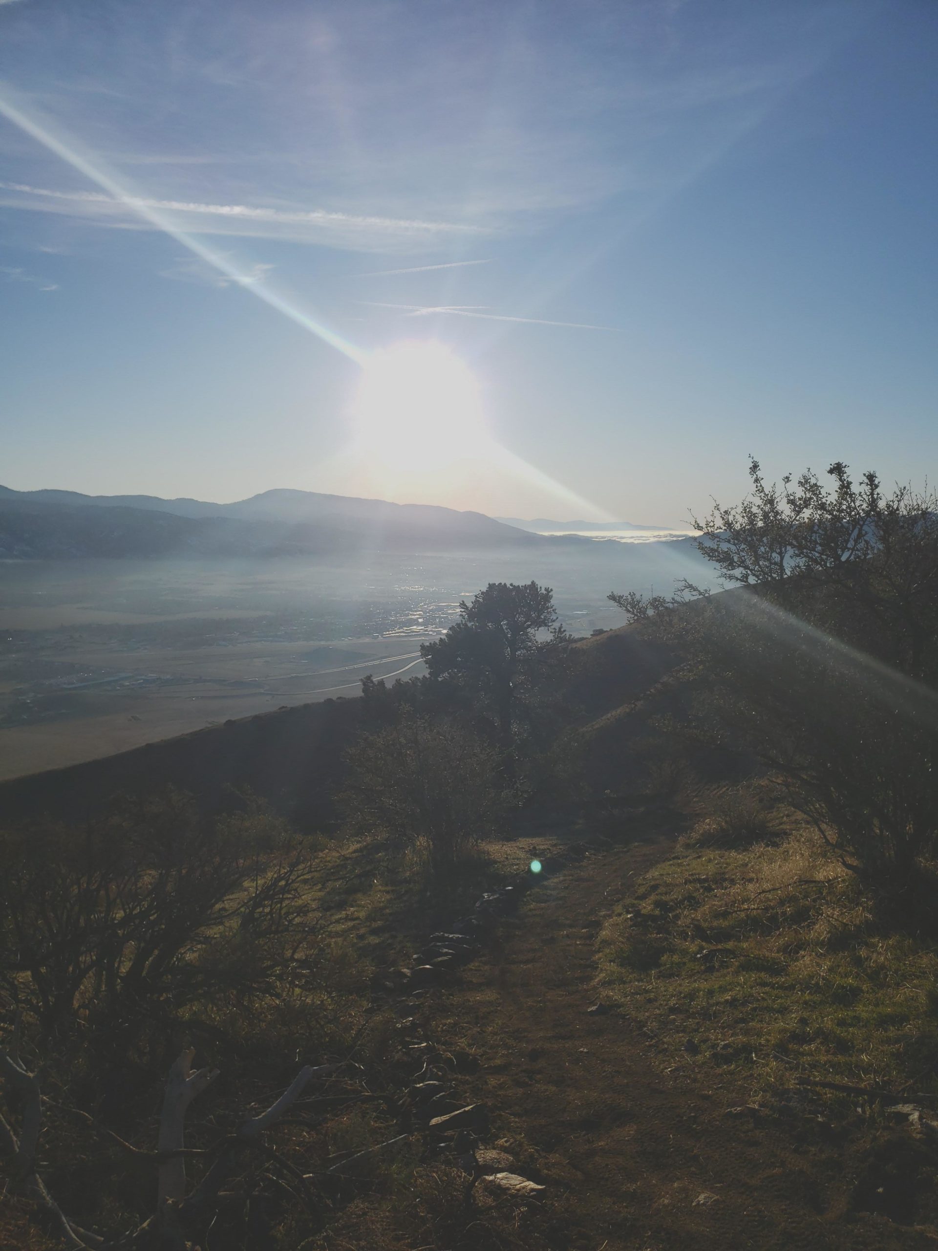 A scenic view of a valley illuminated by the rising sun, with mountains in the background and a clear blue sky. A winding path leads through the foreground, bordered by shrubs and rocks. Soft light beams radiate from the sun, creating a serene atmosphere. TMTA Lehigh trails mountain bike trail.