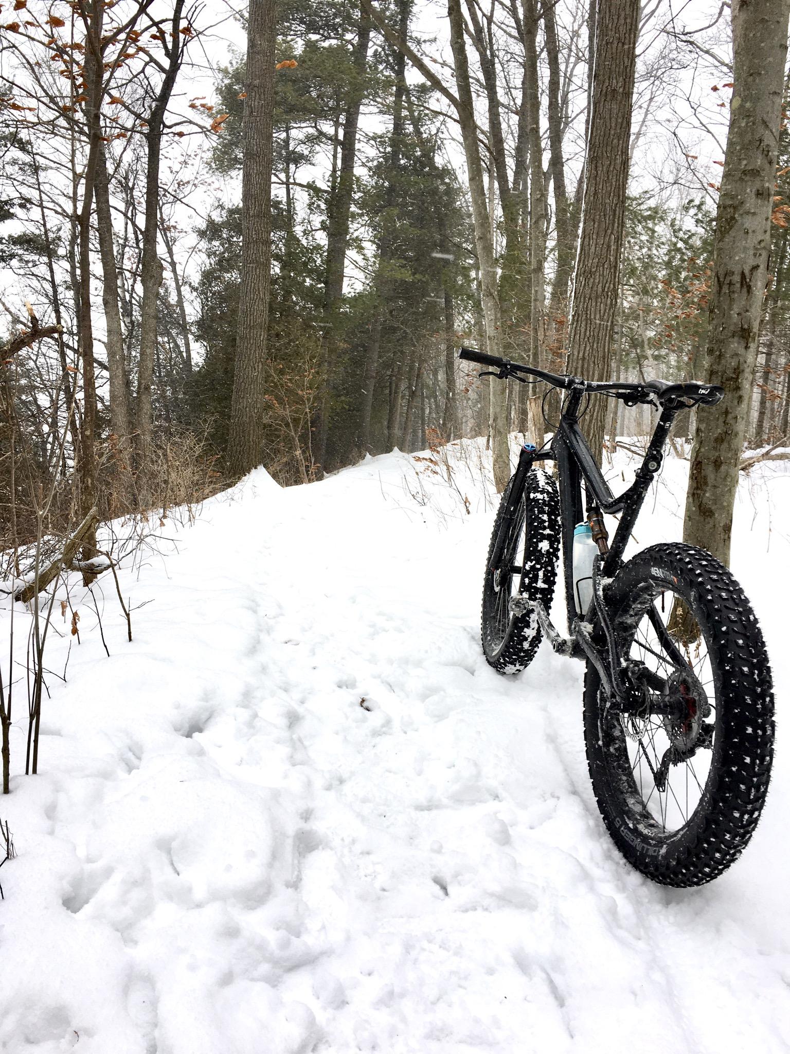 A black fat bike stands on a snow-covered trail in a forest, surrounded by tall trees. Snowflakes can be seen falling gently, creating a serene winter atmosphere. The bike's large tires are designed for traction in snowy conditions. Komoka Trails mountain bike trail.
