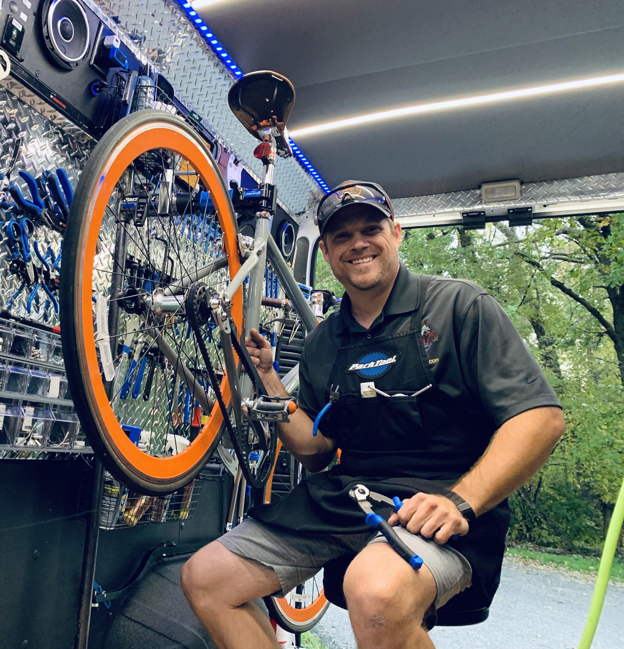 A smiling mechanic working on a bicycle in a mobile workshop. The bike has bright orange rims and is mounted on a stand, surrounded by various tools organized on the wall behind him. The setting includes green trees outside the vehicle, enhancing the outdoor atmosphere.