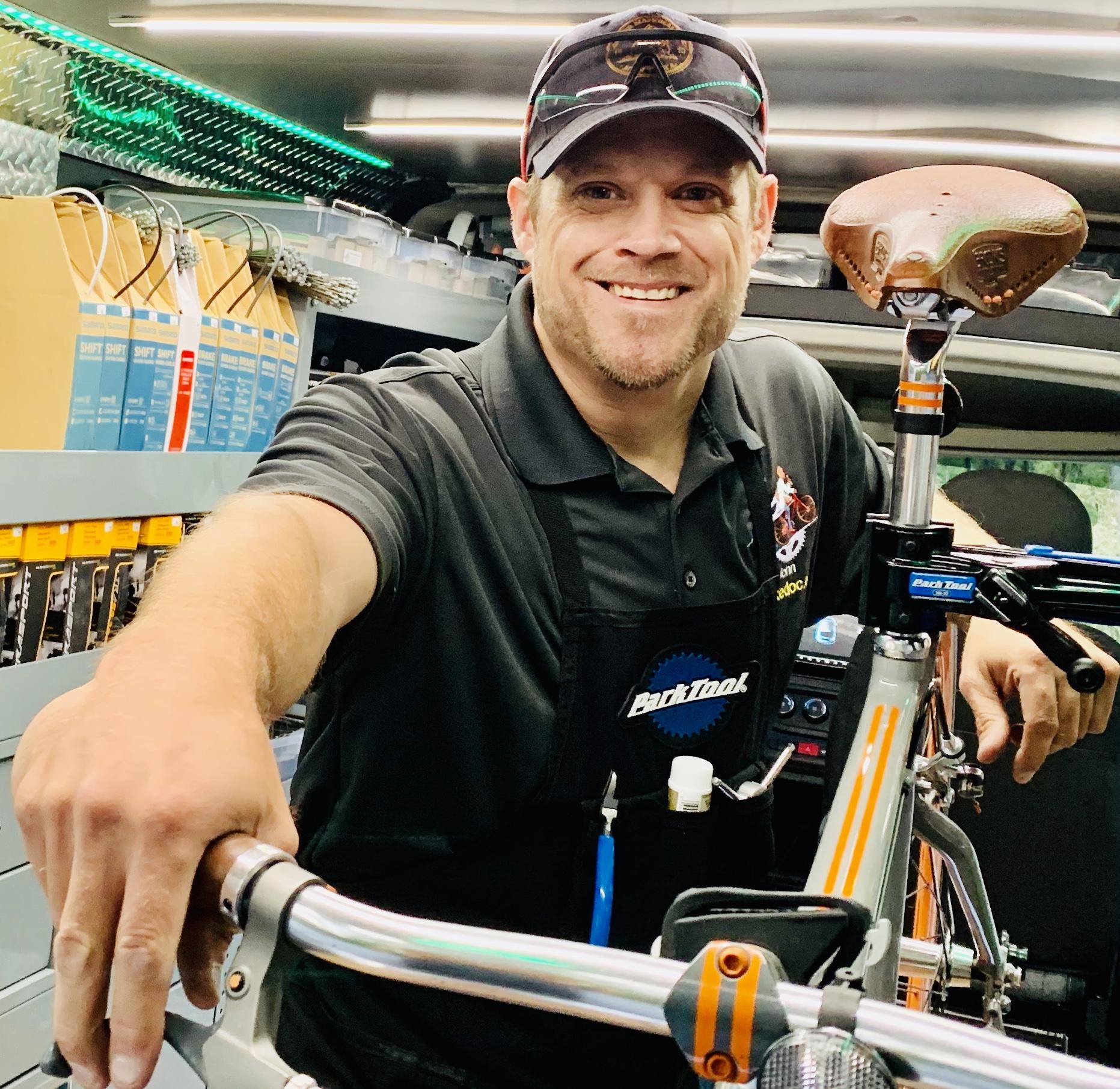 A smiling bike mechanic in an apron stands in a well-equipped workshop, leaning on a bicycle frame with tools and bike accessories visible in the background.