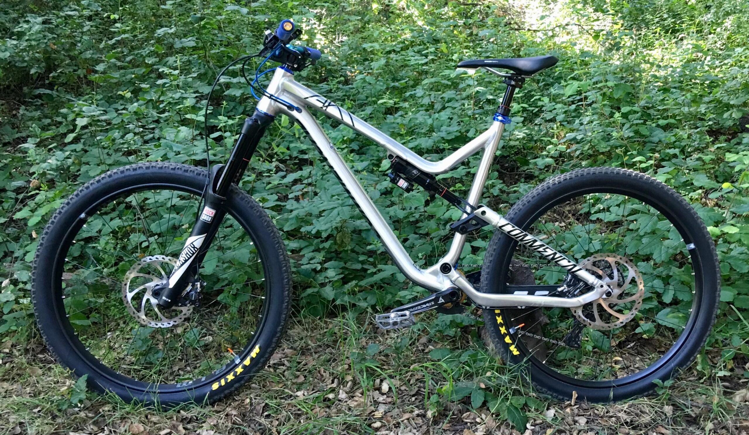 Commencal Meta AM V4: A mountain bike positioned on a trail surrounded by green foliage. The bike features a silver frame, thick tires, and a front suspension fork. The handlebars have blue accents, and the wheels are equipped with disc brakes.