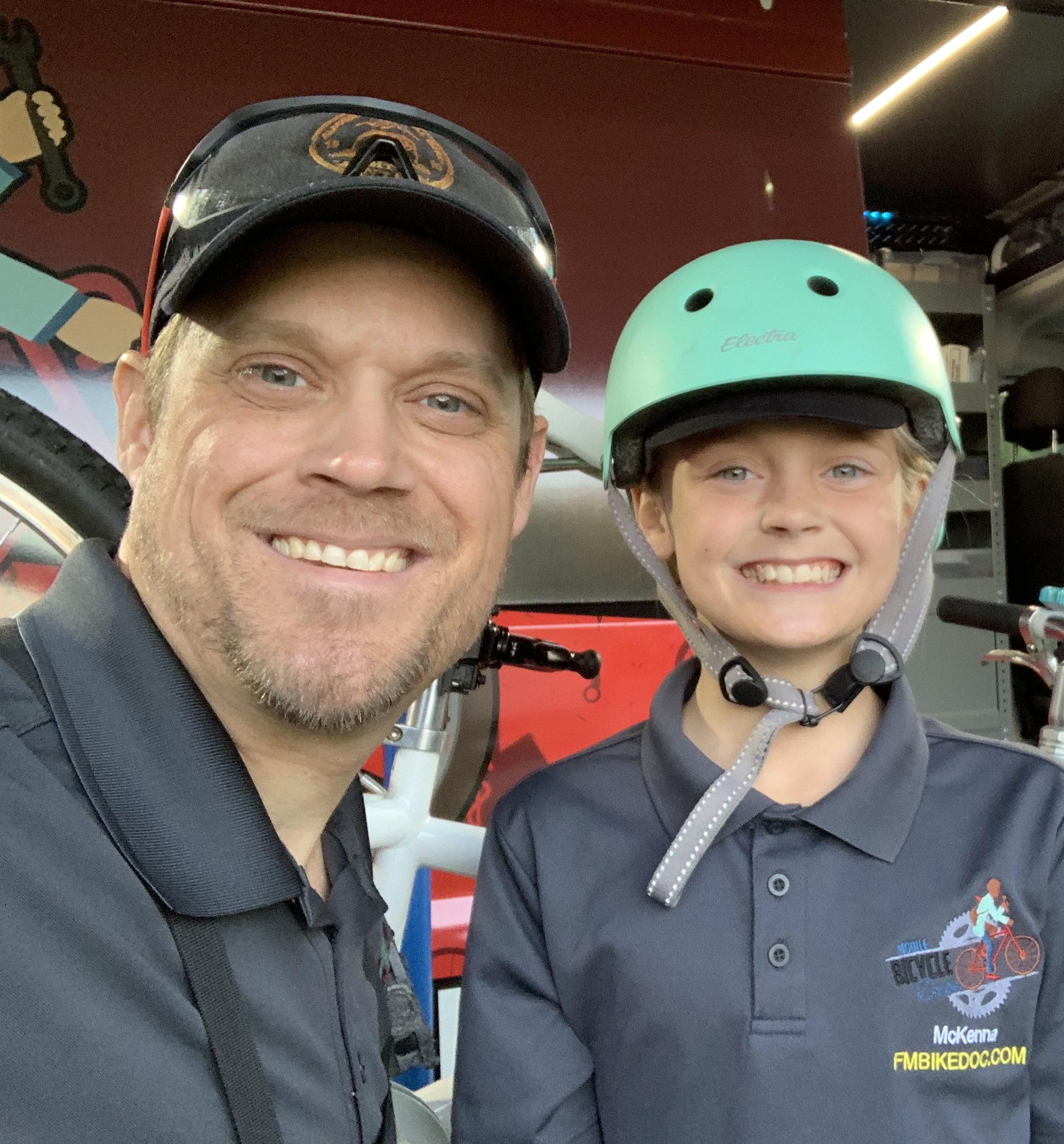 Two smiling individuals pose for a selfie against a backdrop featuring bicycle-themed decorations. A man wearing a baseball cap and a collared shirt stands beside a child wearing a light green helmet and a dark shirt. Both appear happy and engaged, with a bicycle partially visible in the background.