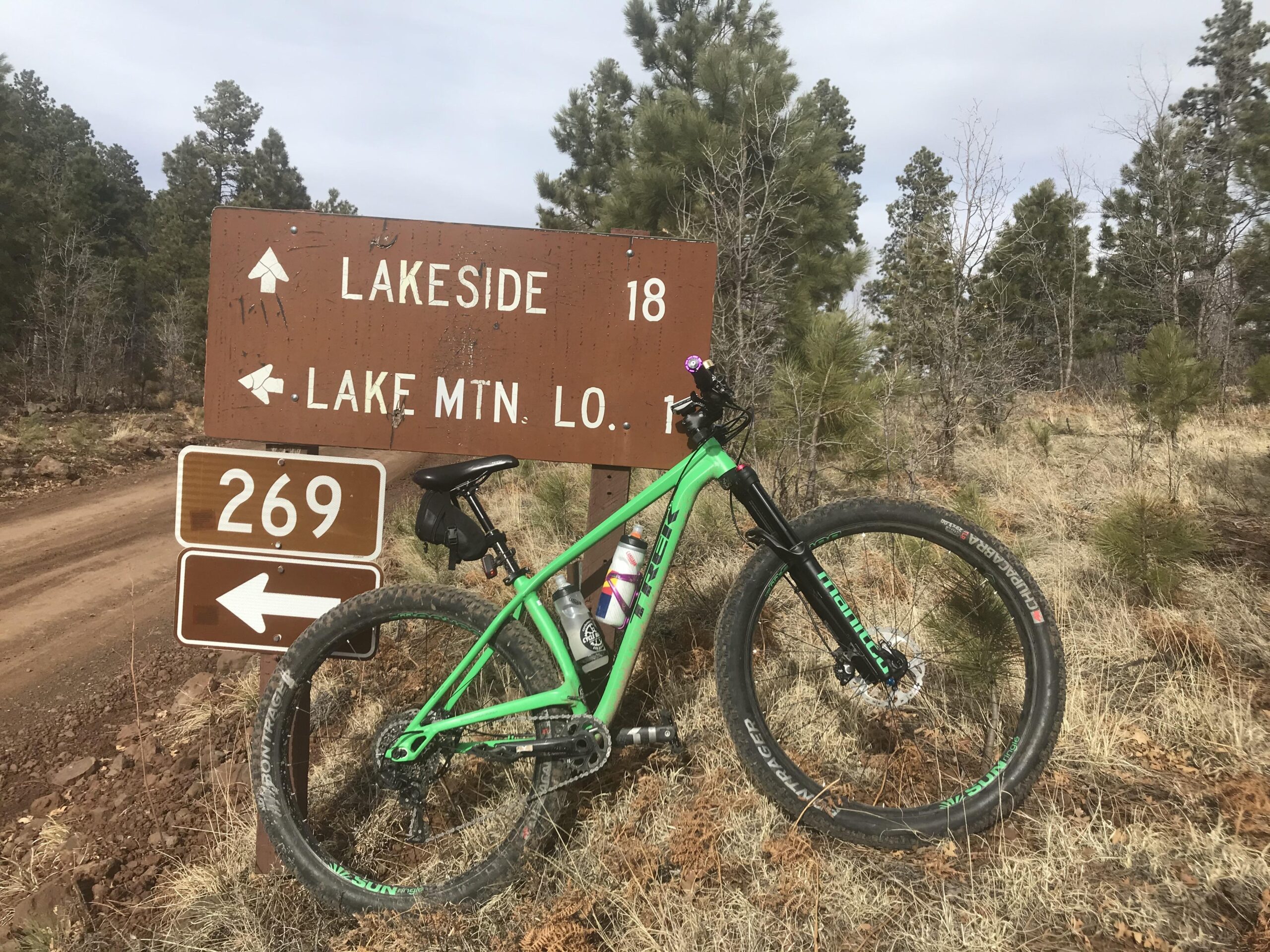 Trek Stache 8: A green mountain bike leaning against a wooden trail sign indicating directions to Lakeside and Lake Mountain Loop, with a nearby road sign marked "269." The background features a forested area with scattered trees and dry grass, under a cloudy sky.
