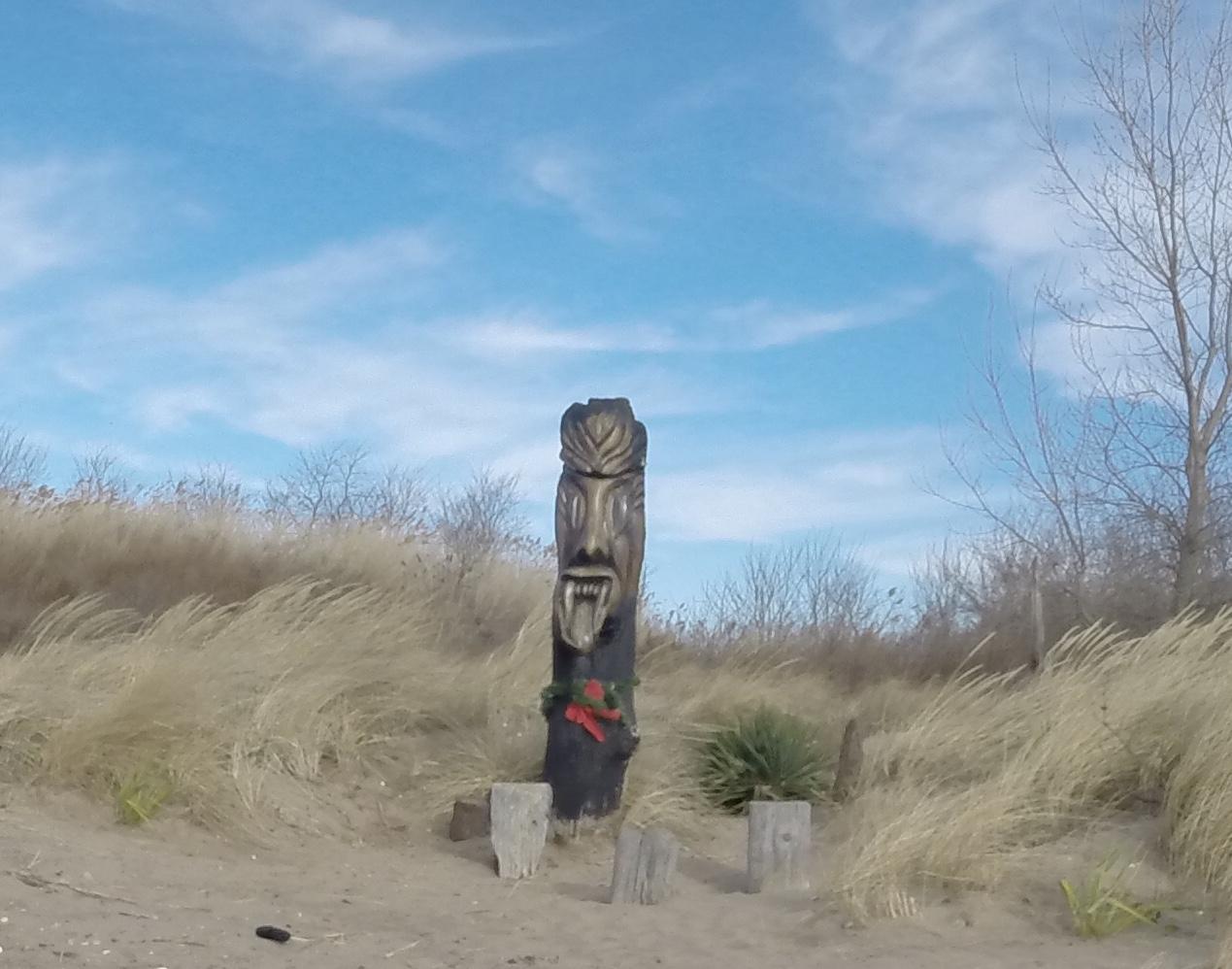 A wooden totem pole with a carved face, adorned with a red ribbon and greenery, situated on a sandy beach surrounded by tall grass under a blue sky with wispy clouds. Beach Long Pond Area mountain bike trail.