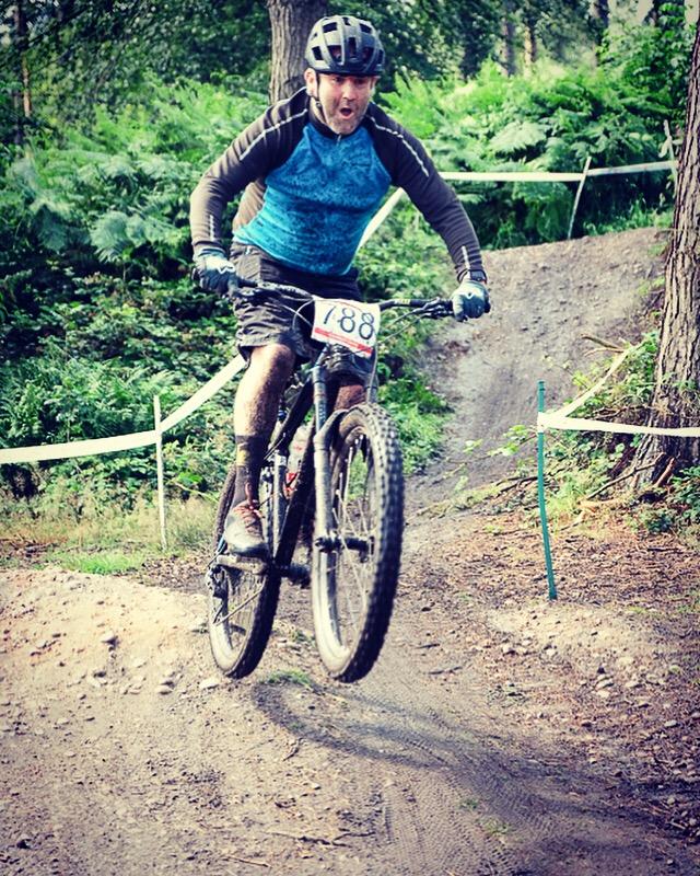 Vitus Sentier 29 (1x10): A mountain biker wearing a blue and black jersey and a helmet is captured mid-jump on a dirt trail surrounded by greenery. The cyclist has a race number, 188, displayed on their bike. Mud splatters are visible on their legs and bike, indicating an adventurous ride.