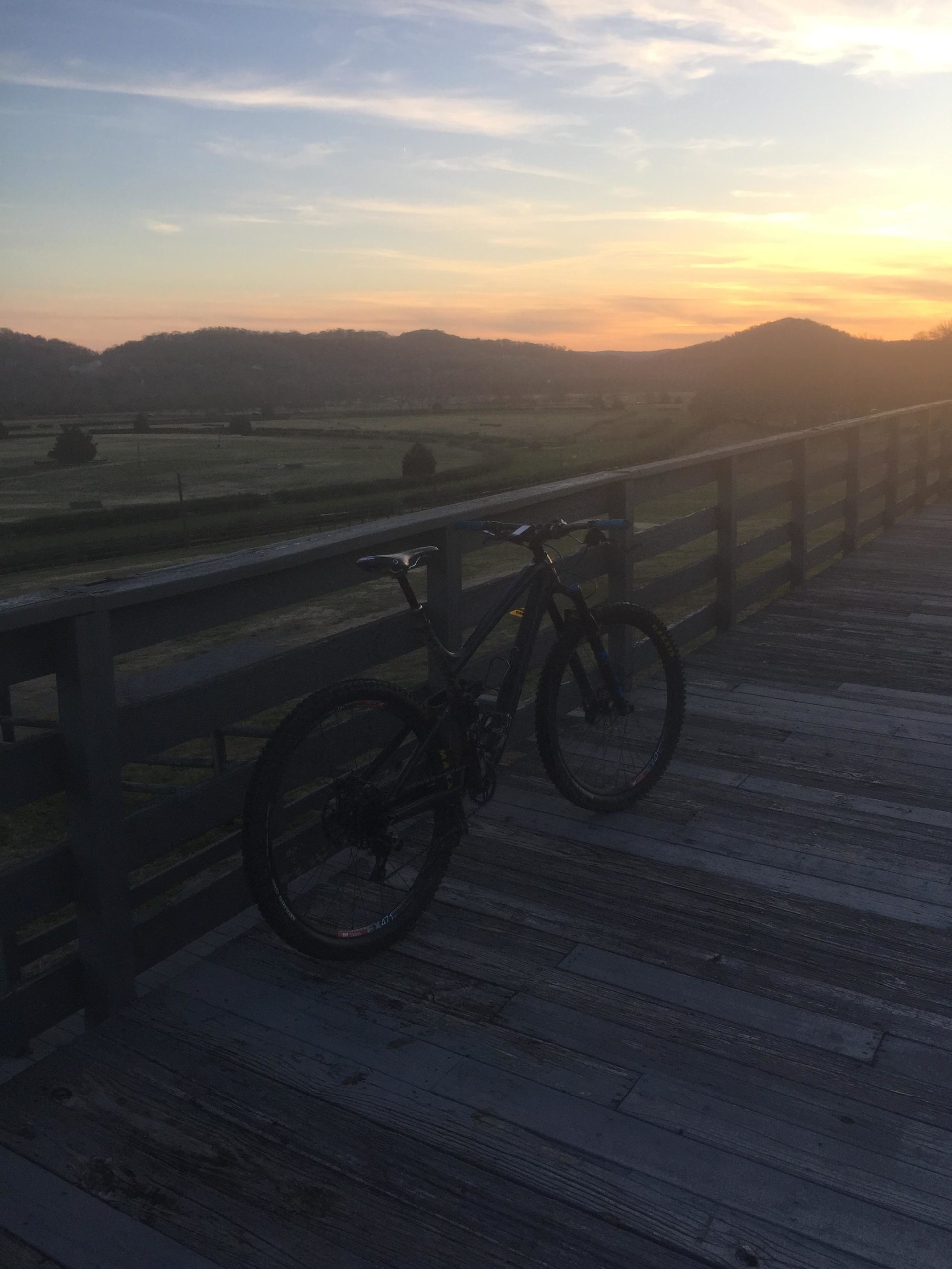 A mountain bike leans against a wooden railing at sunset, overlooking a serene landscape of rolling hills and fields. The sky is painted with soft hues of orange and blue, creating a tranquil outdoor scene. Percy Warner Mountain Bike Trails mountain bike trail.