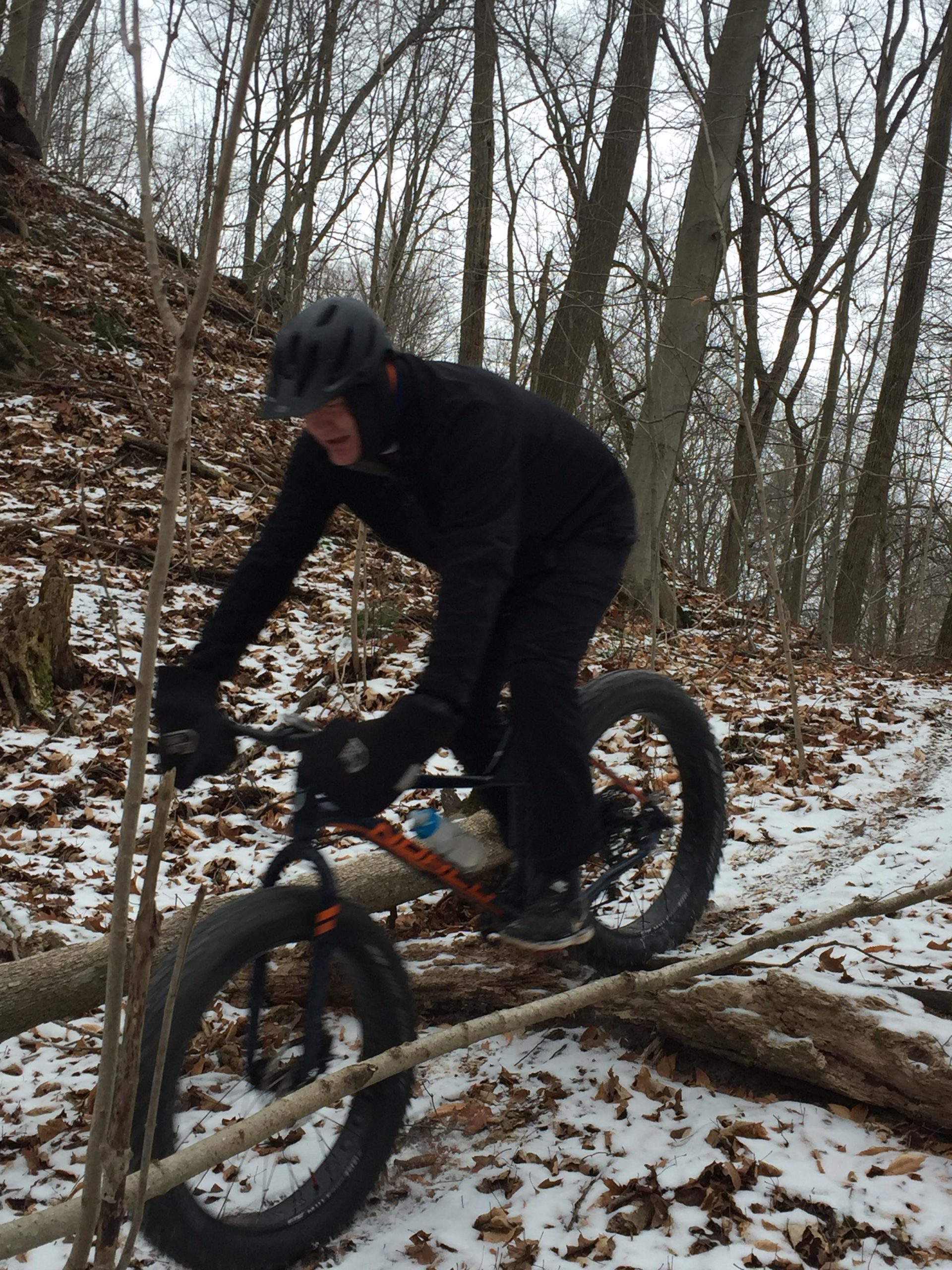 A person wearing a black helmet and winter clothing is riding a fat bike over a fallen log on a snowy trail surrounded by bare trees. The ground is covered with snow and fallen leaves, creating a wintry scene in a forested area. Swance Drain mountain bike trail.
