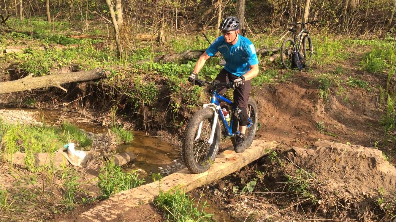 A mountain biker in a blue shirt navigates a narrow wooden bridge over a small stream, surrounded by greenery and trees in a wooded area. Swance Drain mountain bike trail.