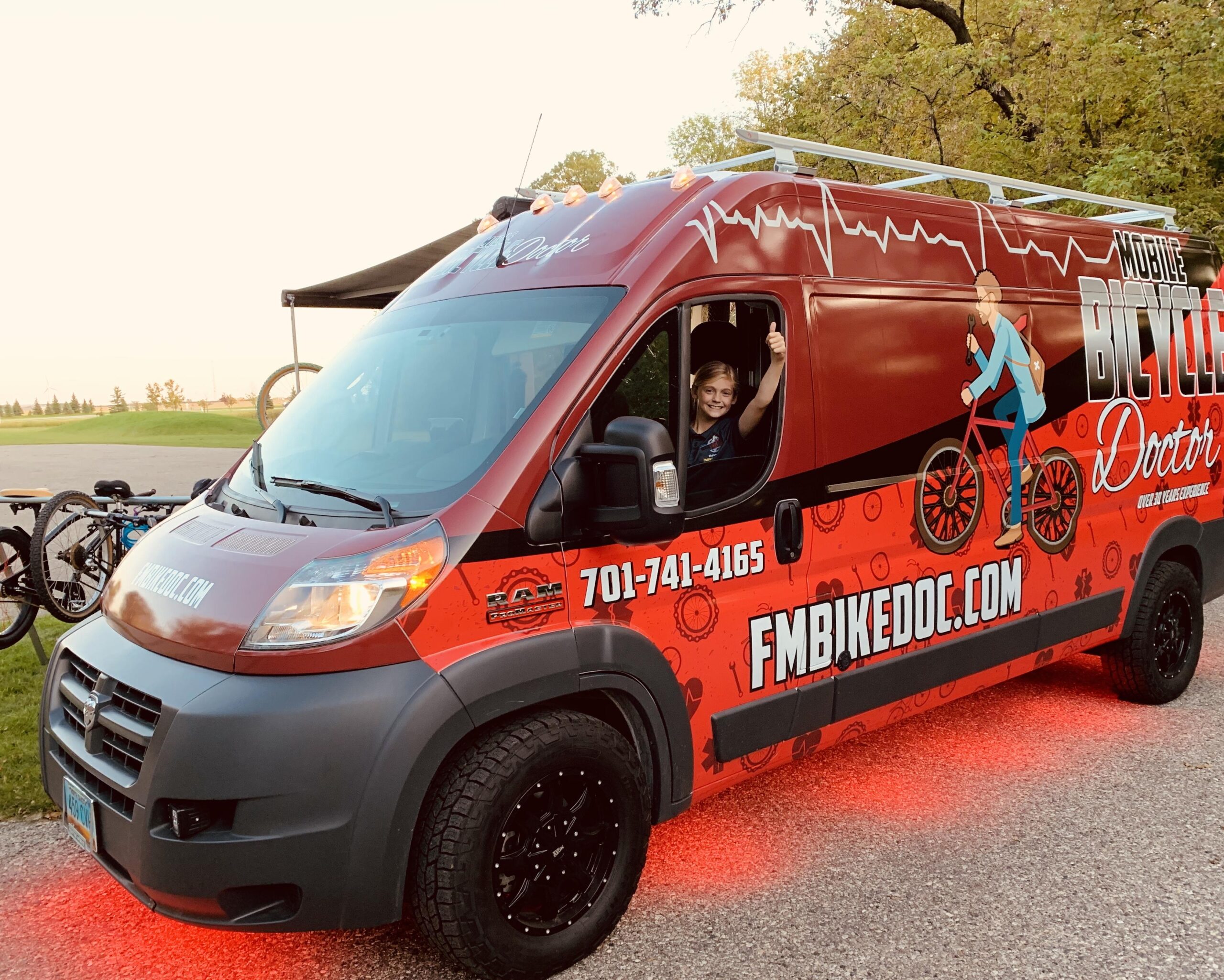 A red and gray van labeled "Mobile Bicycle Doctor" with images of bicycles and gears on its sides. A smiling person inside the driver's seat is giving a thumbs-up. The vehicle is parked on a road with bicycles in the background, and the ground under the van is illuminated with red light, surrounded by greenery.