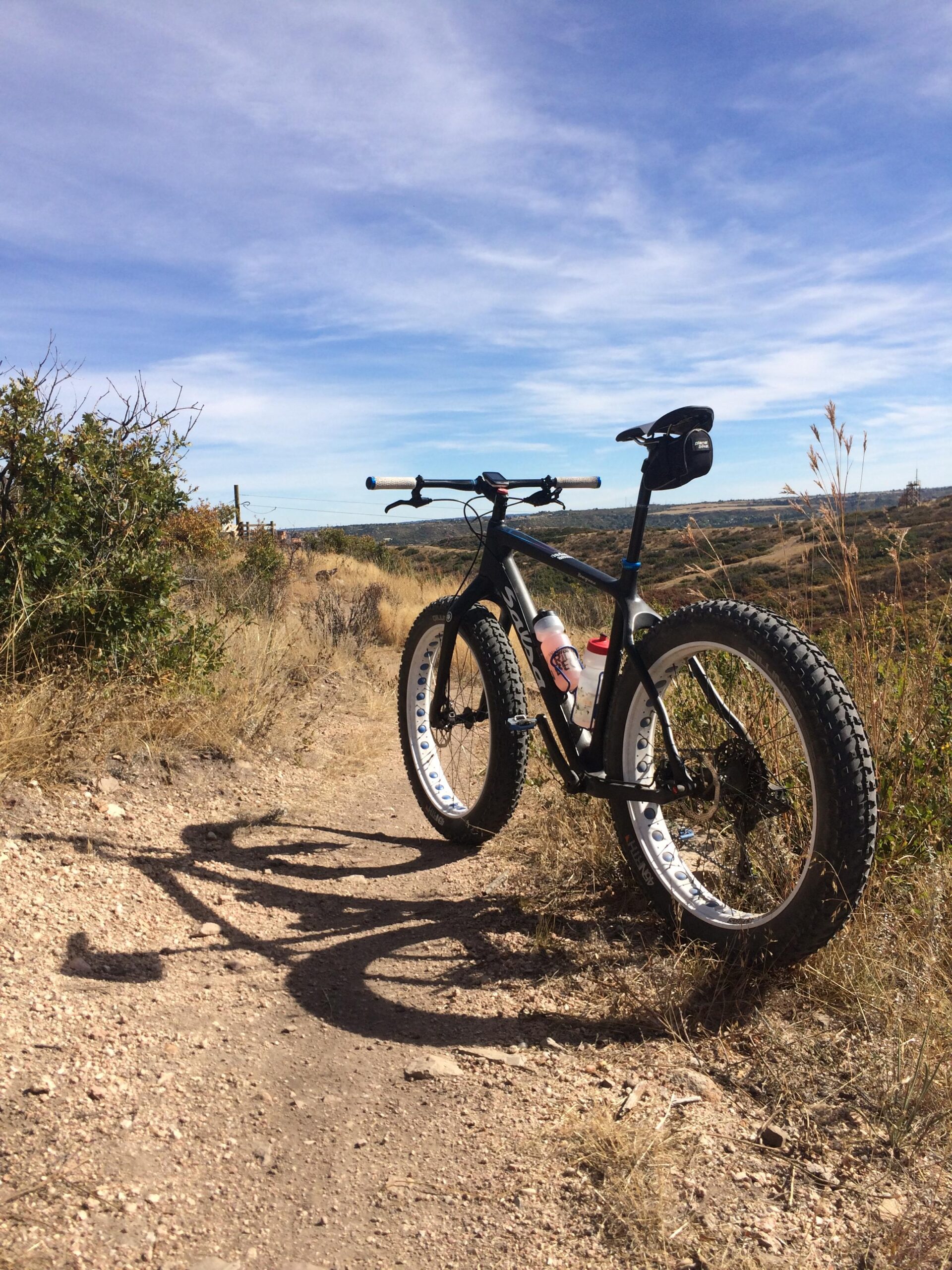 Salsa Beargrease: A fat tire mountain bike parked on a dirt trail surrounded by dry grass and shrubs, with a clear blue sky overhead. The bike features wide tires and a water bottle holder, casting a long shadow on the ground.