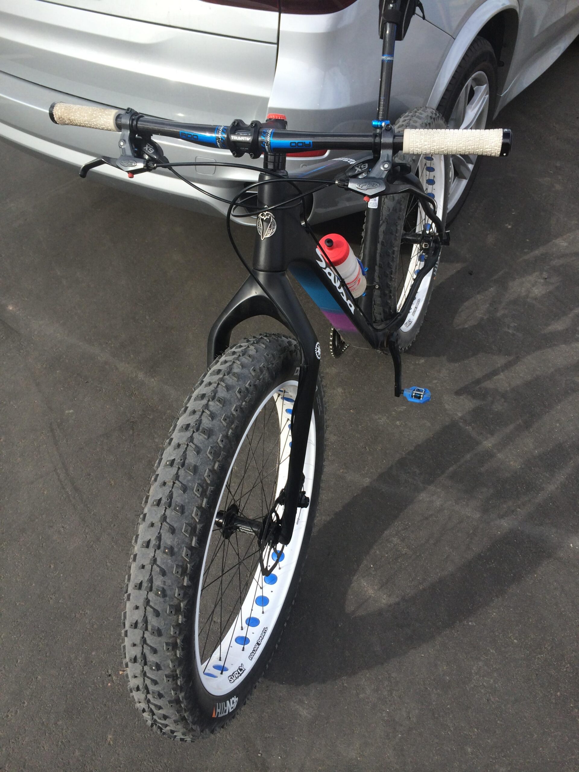 Salsa Beargrease: Alt text: A close-up view of a black mountain bike with wide tires, showing the handlebars, brake levers, and a water bottle mounted on the frame. In the background, a silver SUV is parked on an asphalt surface.