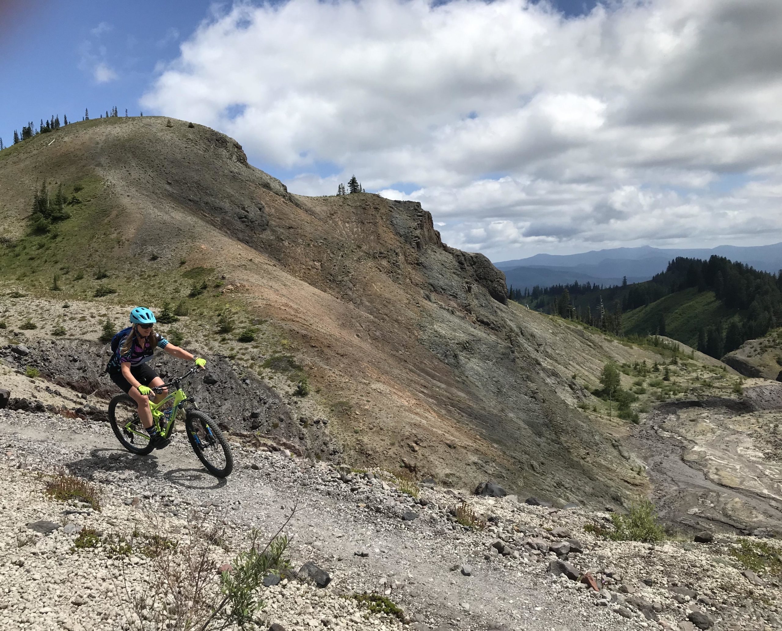 A child riding a mountain bike down a rocky trail on a hillside, surrounded by lush green trees and rugged terrain under a partly cloudy sky. Ape Canyon#234, Abraham#216d, Smith Creek#225 Trails mountain bike trail.