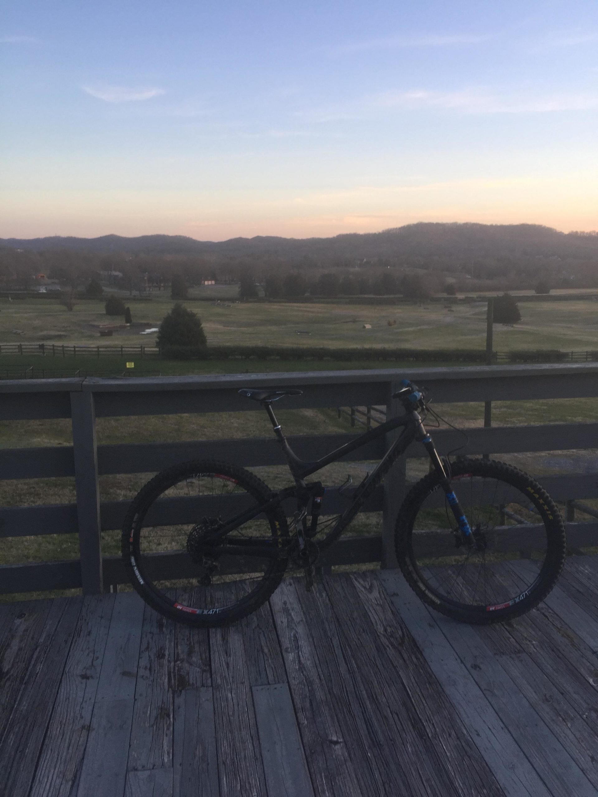 A mountain bike resting on a wooden deck with a scenic view of rolling hills and a field in the background during sunset. Percy Warner Mountain Bike Trails mountain bike trail.