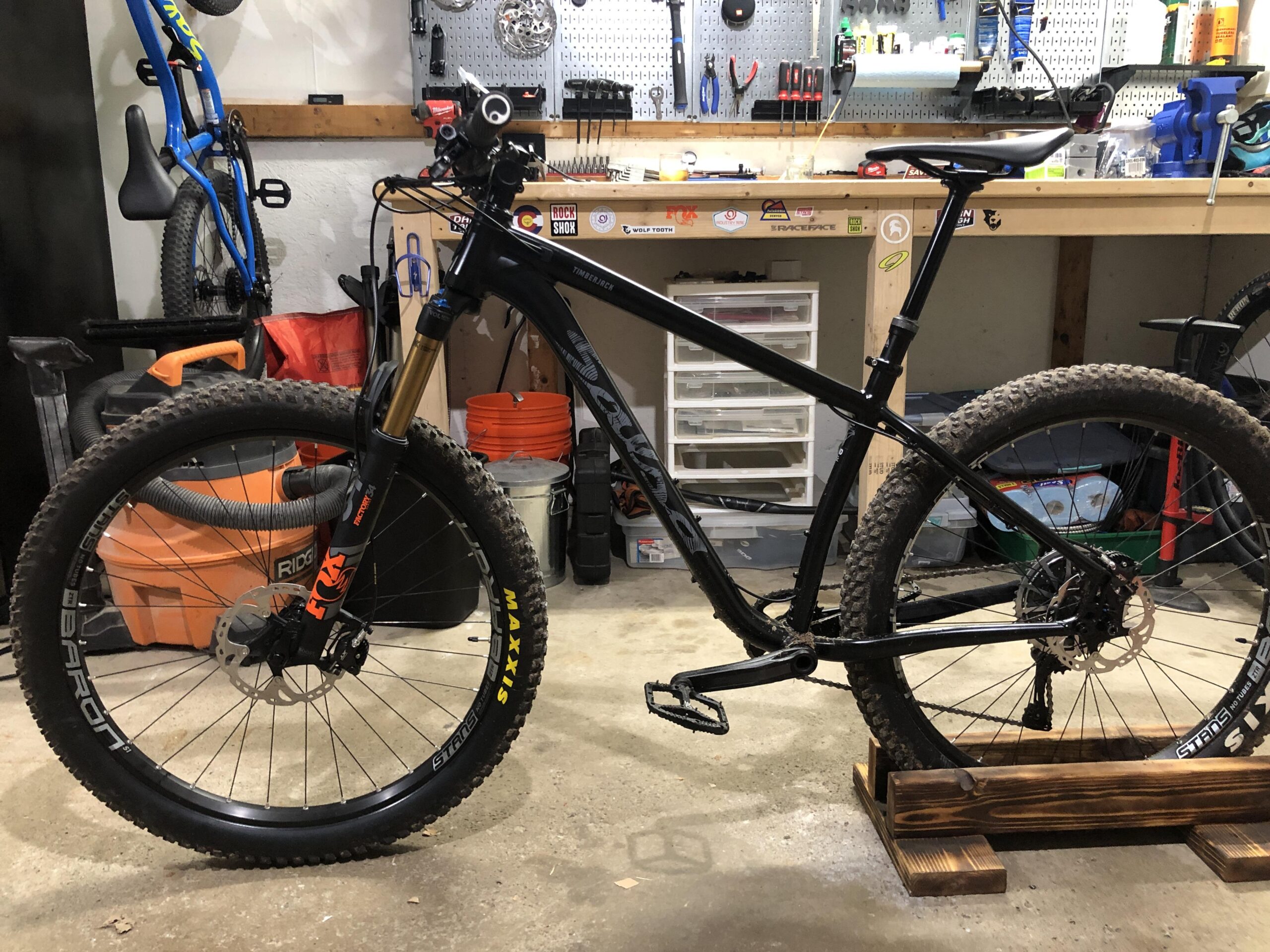 Salsa Timberjack: A black mountain bike with thick tires and a modern frame stands upright on a wooden bike stand in a garage. The bike features front suspension with a gold fork, a dropper seat post, and has a rear cassette visible. Various tools and bike accessories are organized on a workbench in the background, along with a blue bike hanging on the wall.