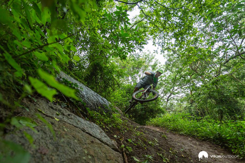 A mountain biker performing a jump on a rugged trail, surrounded by lush greenery and trees. The bike is airborne over a rocky section of the path, showcasing the dynamic action of the sport. Giba Green Route mountain bike trail.
