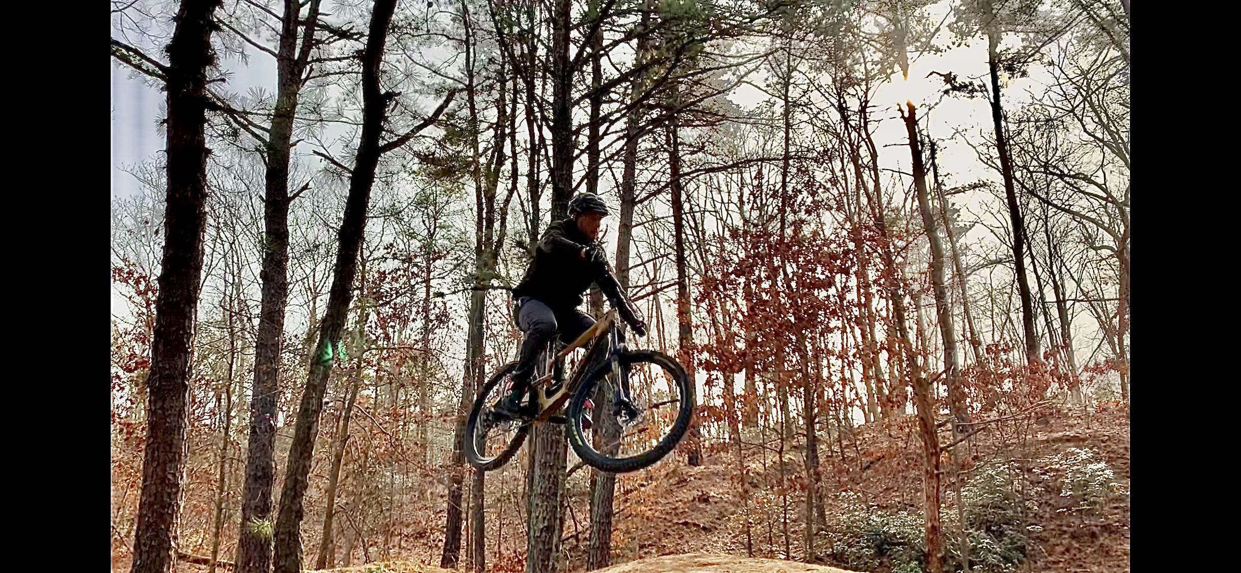 Santa Cruz Nomad: A mountain biker performing a jump on a trail surrounded by trees, with autumn foliage in the background. The cyclist is wearing a helmet and riding a mountain bike, capturing a moment of action and excitement in nature.