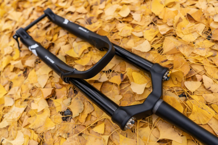 A close-up view of a black bicycle fork resting on a bed of yellow autumn leaves. The fork features smooth lines and modern design elements, highlighting its suspension components and attachment points. The vibrant color of the leaves contrasts with the dark finish of the fork, creating a visually appealing autumnal scene.