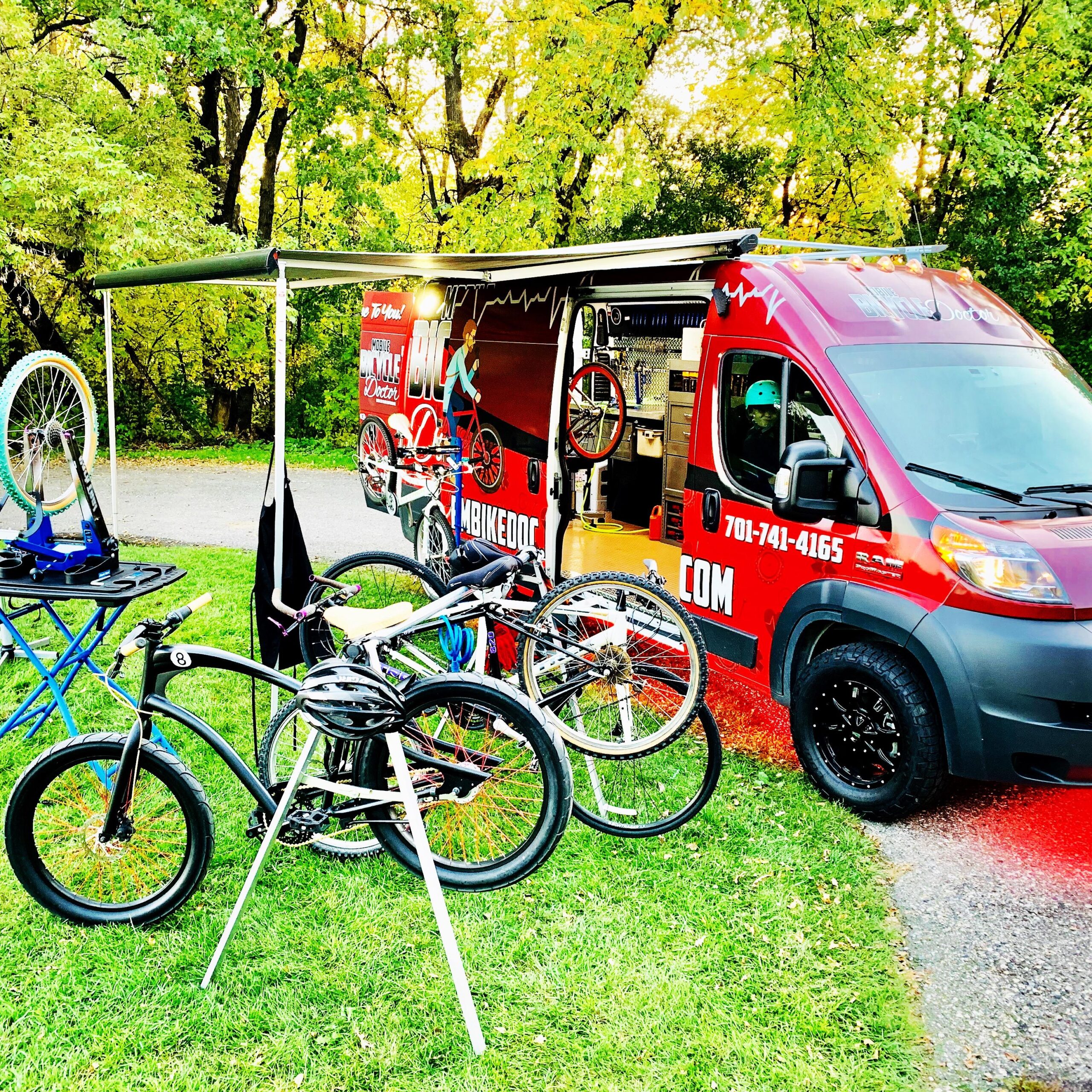A vibrant mobile bike repair service parked in a scenic area. The van, featuring eye-catching graphics of bicycles and contact information, is set up with an awning. Several bikes are displayed nearby, with some on repair stands, showcasing a variety of styles. The surrounding landscape features lush greenery, creating a welcoming atmosphere for cycling enthusiasts.