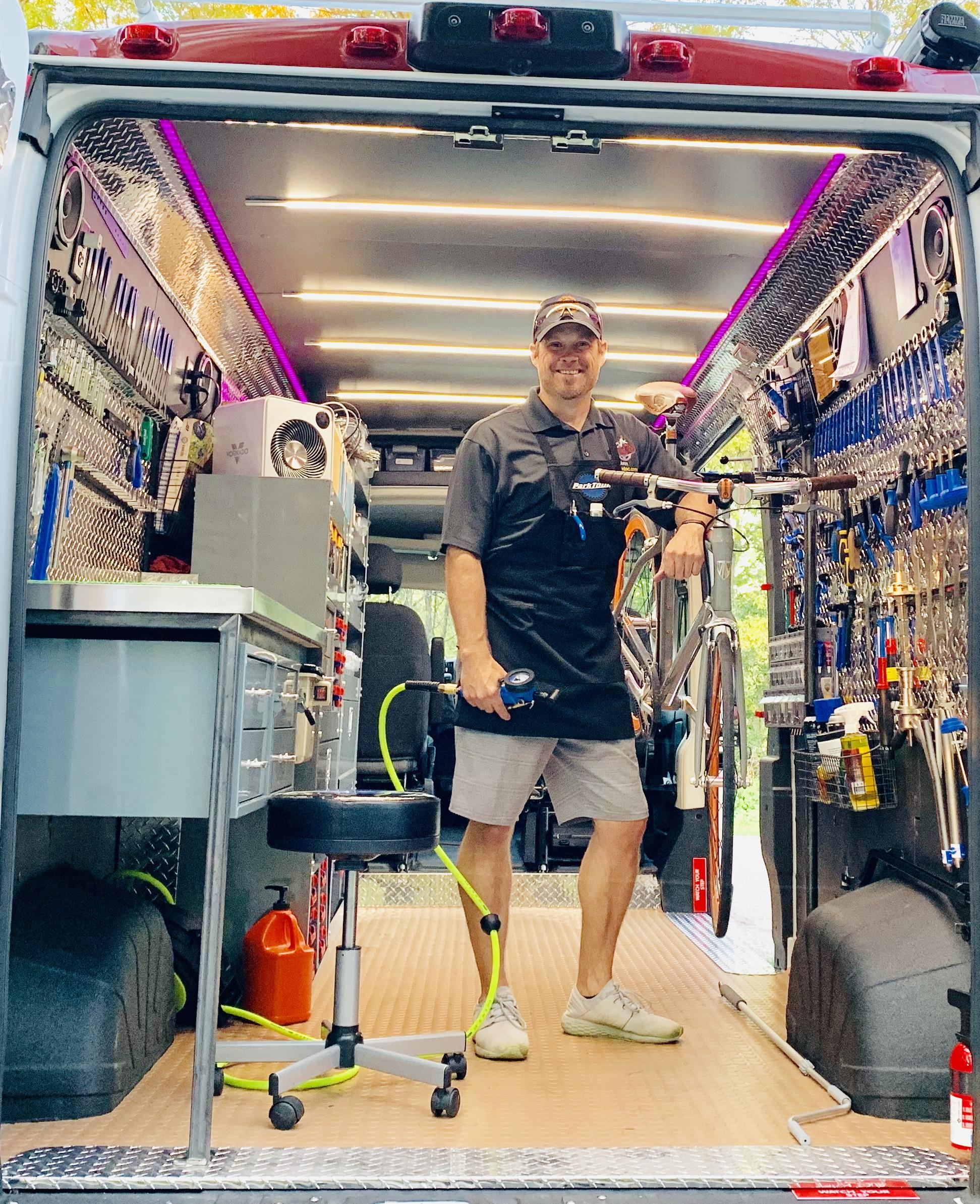 A man stands inside a well-equipped mobile workshop, smiling and posing with a bicycle. The van's interior is organized with various tools and equipment mounted on the walls, and a workbench is visible to the side. Bright LED lights illuminate the space, creating a professional and inviting atmosphere.