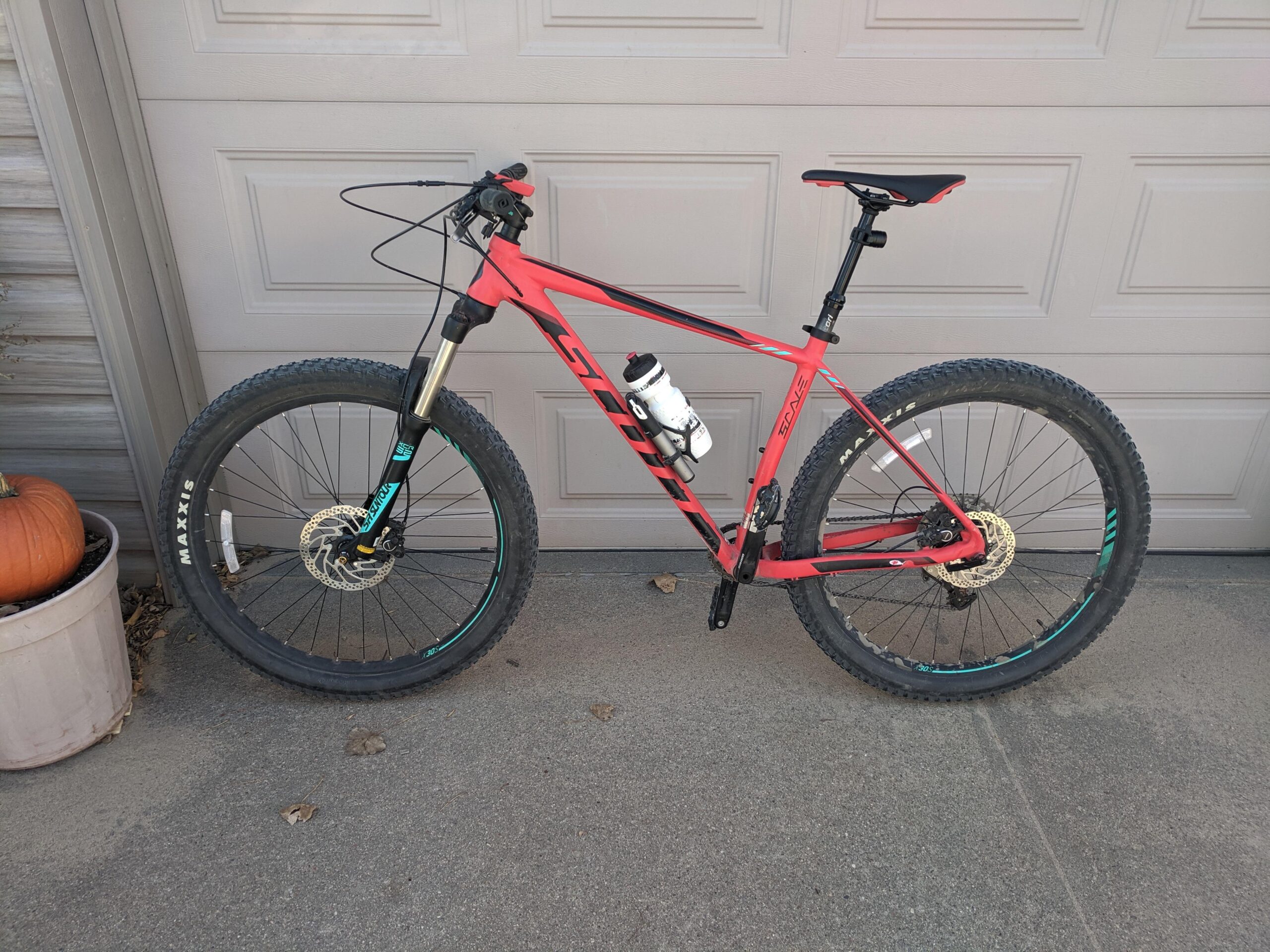 Scott Scale 730: A red mountain bike with thick tires, a water bottle attached to the frame, and a dropper seat post, leaning against a garage door. A pumpkin is visible in a planter nearby, with a paved surface and some fallen leaves on the ground.