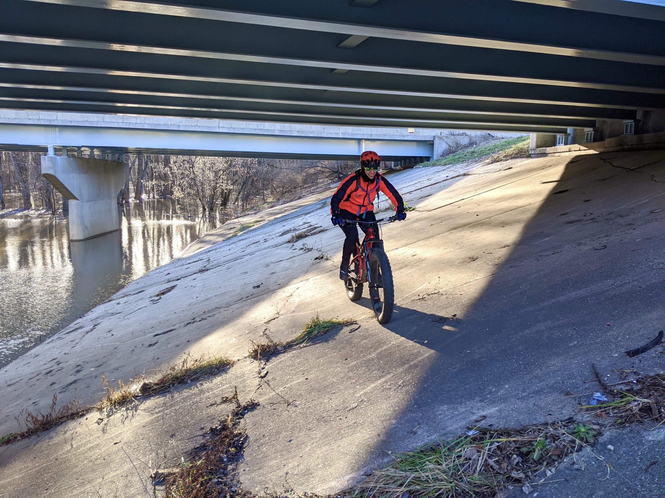 A person wearing a bright orange jacket and a helmet rides a fat bike along a sloped concrete bank under a bridge. The scene includes a calm river on one side and trees in the background. Sunlight creates contrasting shadows on the surface. Wabashiki Trail mountain bike trail.