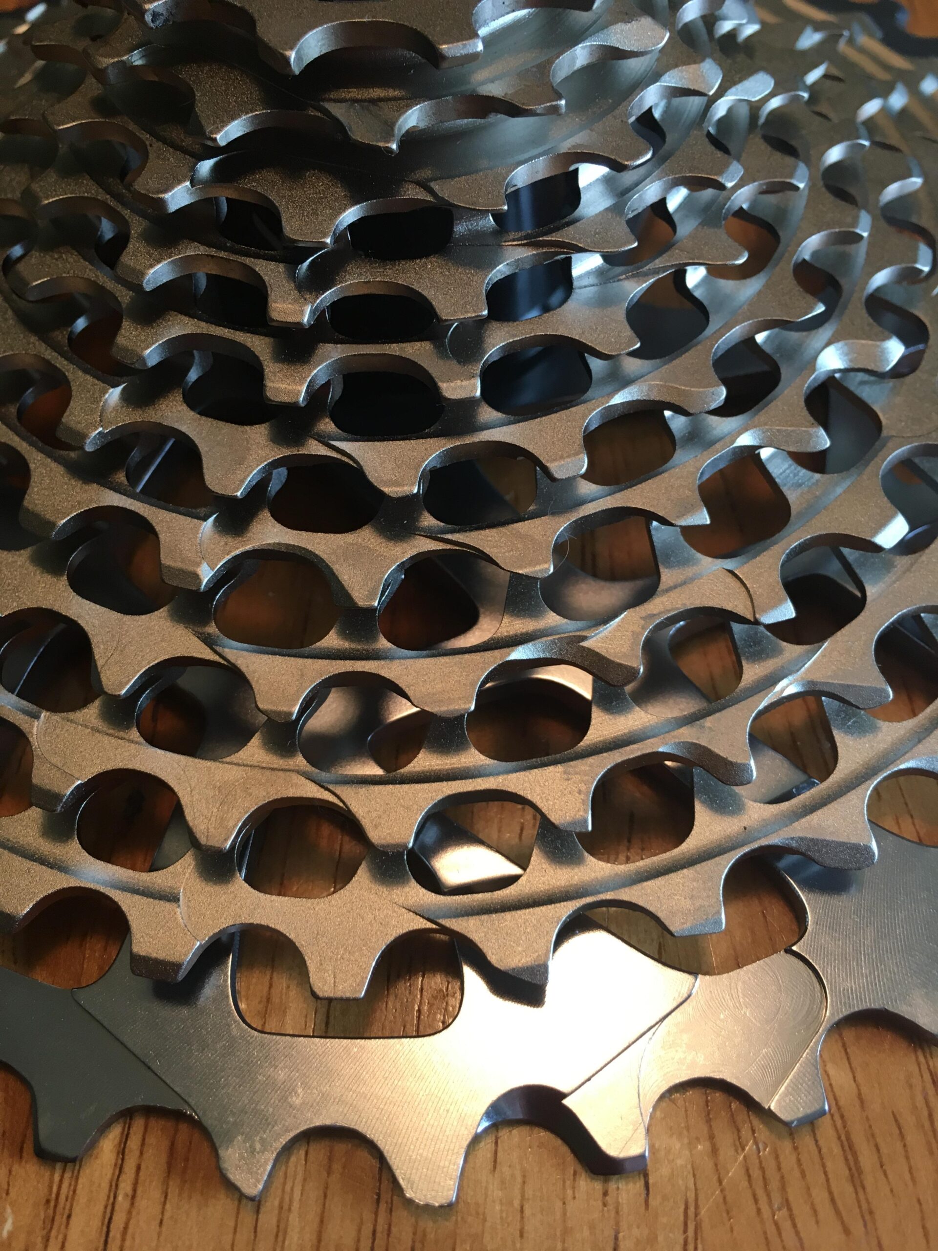 A close-up image of stacked bicycle cogs, showcasing their metallic surfaces and intricate designs. The cogs are arranged in a circular pattern, highlighting the varying sizes and shapes of the teeth, with a wooden surface visible beneath them.