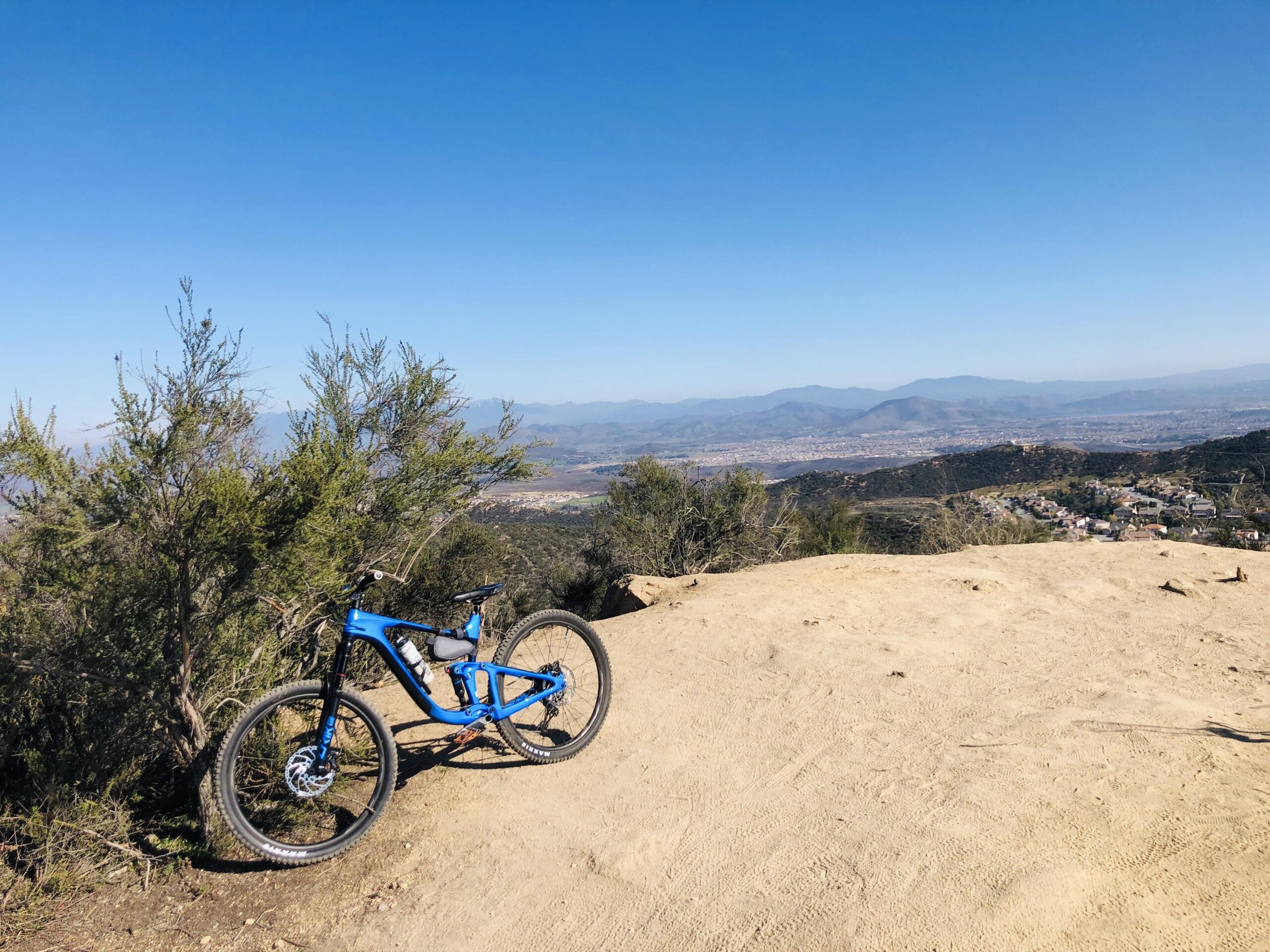 Giant Reign: A blue mountain bike resting on a dirt trail, with a panoramic view of distant mountains and a valley in the background, under a clear blue sky.