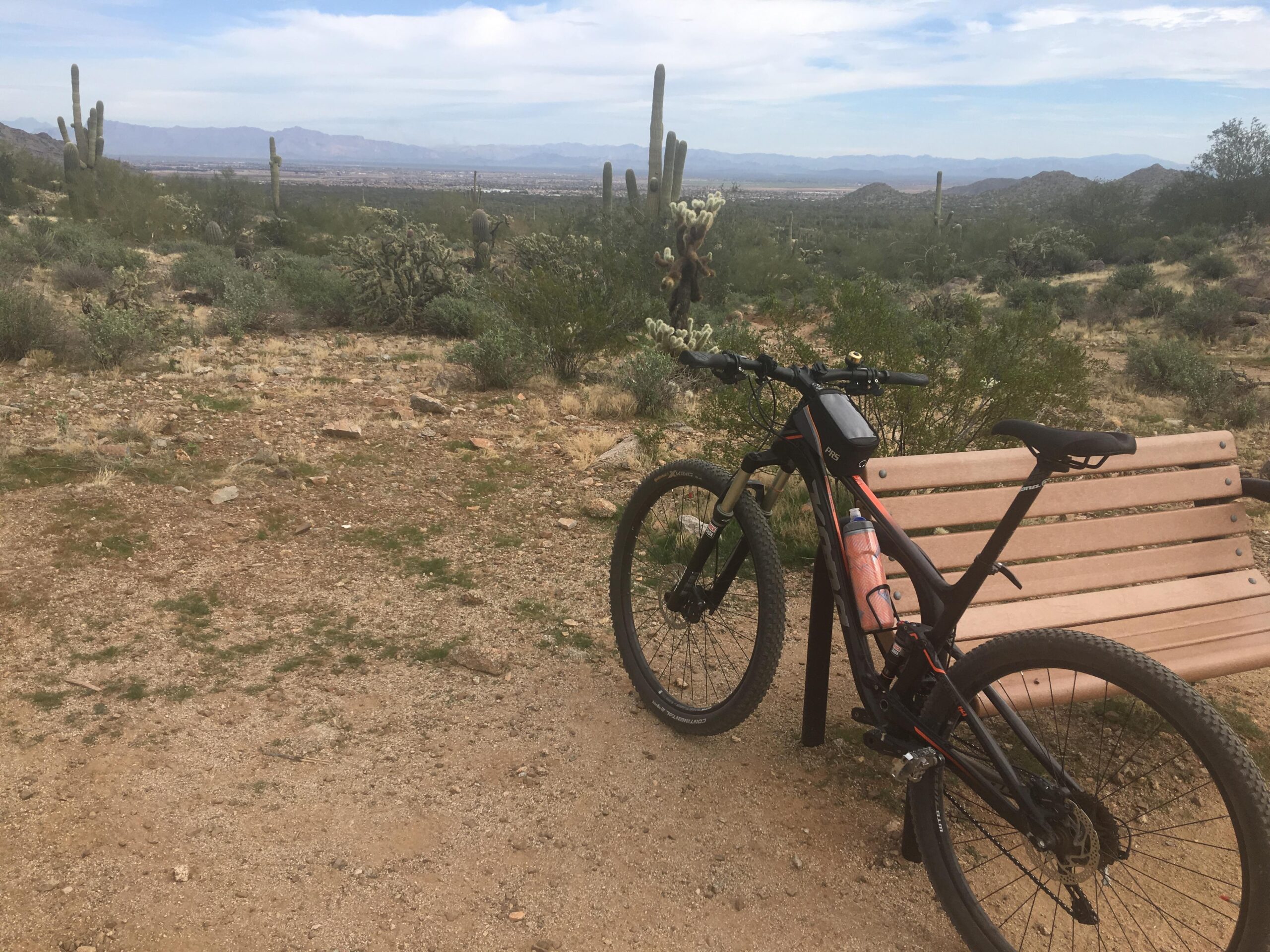 Kona Hei Hei: A black mountain bike parked next to a wooden bench in a desert landscape. The scene features cacti, shrubs, and rocky terrain, with mountains visible in the background under a cloudy sky. A water bottle is attached to the bike.