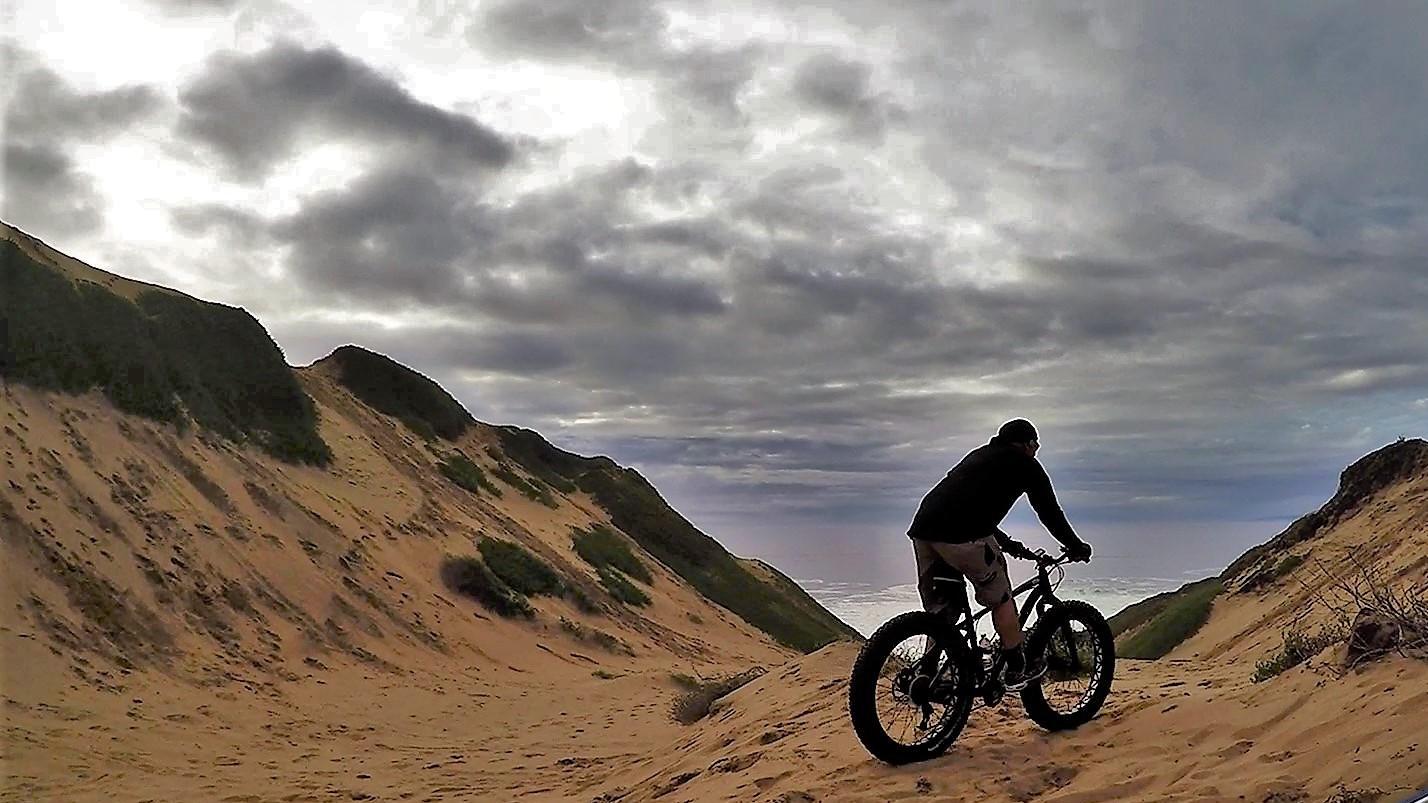 Specialized Fatboy: A person riding a mountain bike on sandy terrain, surrounded by rolling hills and a cloudy sky. The scene captures a sense of adventure and the beauty of outdoor exploration.