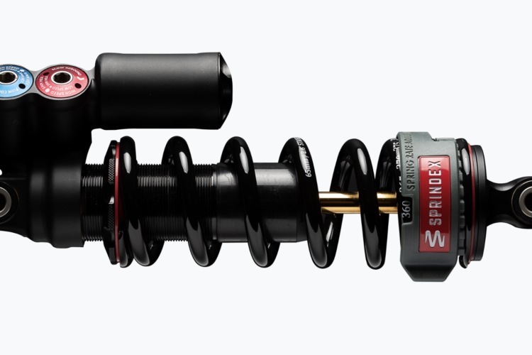 An image of a bicycle shock absorber featuring a black coil spring, an adjustment knob with blue and red markings for high and low speed compression settings, and a gray and red body labeled "SPRINDEX." The shock absorber is shown from a side view, highlighting its mechanical design and components.