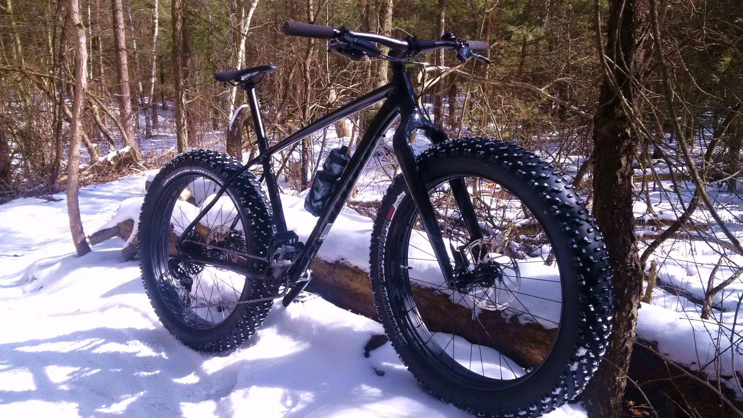 Specialized Fatboy: A black fat bike with wide, studded tires rests on a snowy forest path, surrounded by trees. The bike features a sleek frame and is equipped with a water bottle mounted on the frame. Snow covers the ground, and a fallen log is visible in the foreground, creating a serene winter scene.