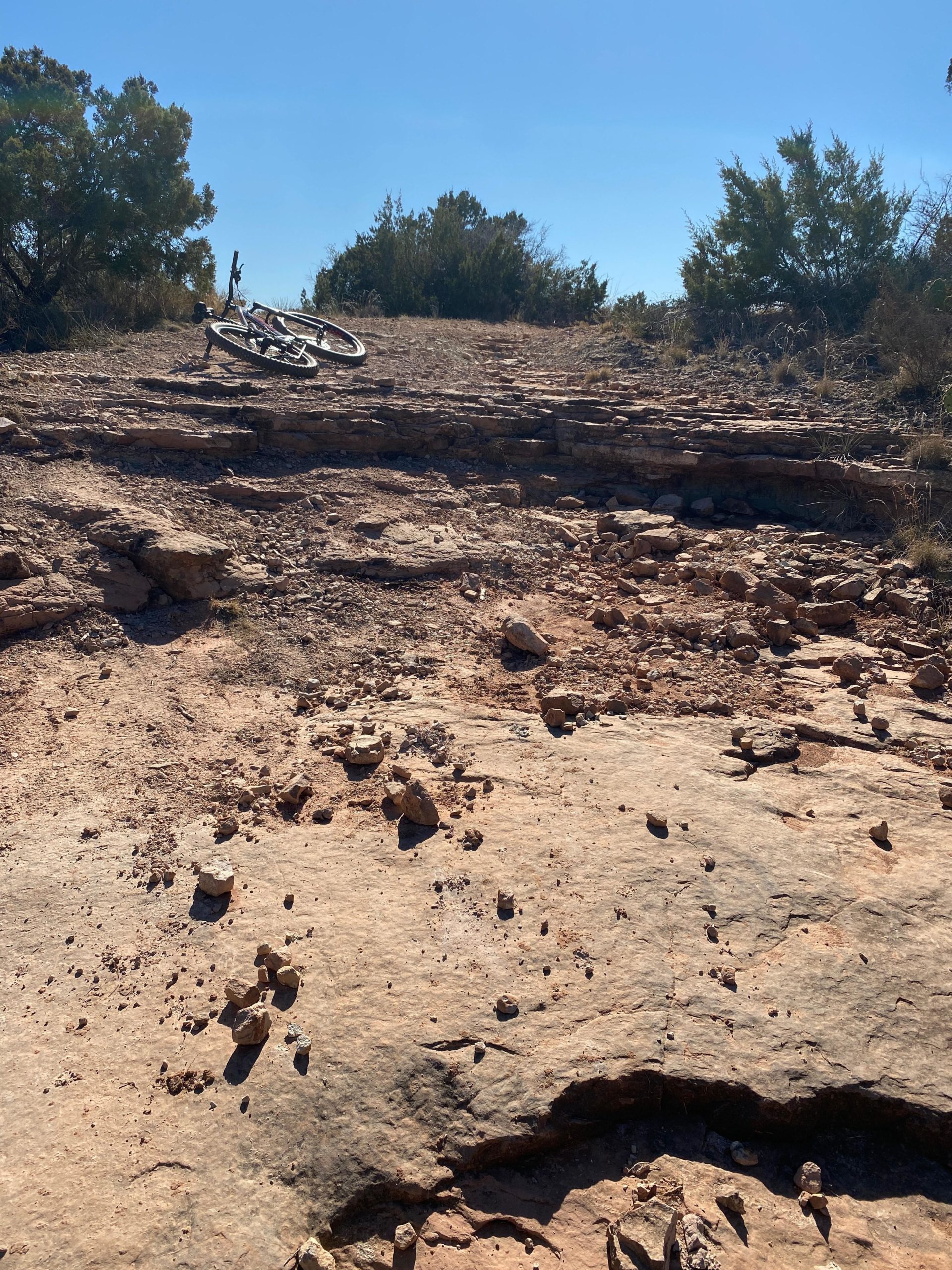 A rocky mountain biking trail with a bicycle lying on its side. The path is uneven and gravelly, surrounded by sparse vegetation under a clear blue sky. Twin Butte Open Space mountain bike trail.