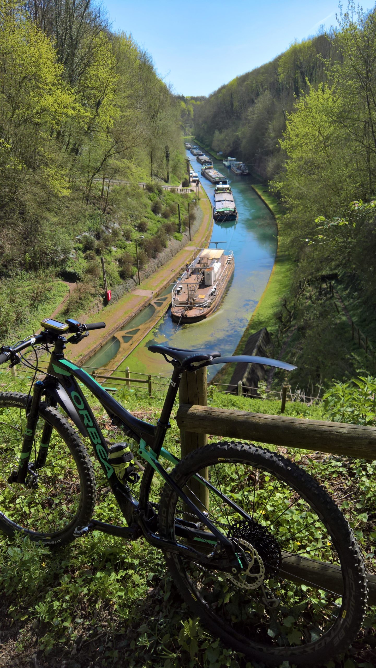 A mountain bike resting on a grassy hillside, overlooking a serene canal lined with trees. Several boats are docked along the waterway, which winds through a lush green landscape under a clear blue sky. Hidenbourg line mountain bike trail.
