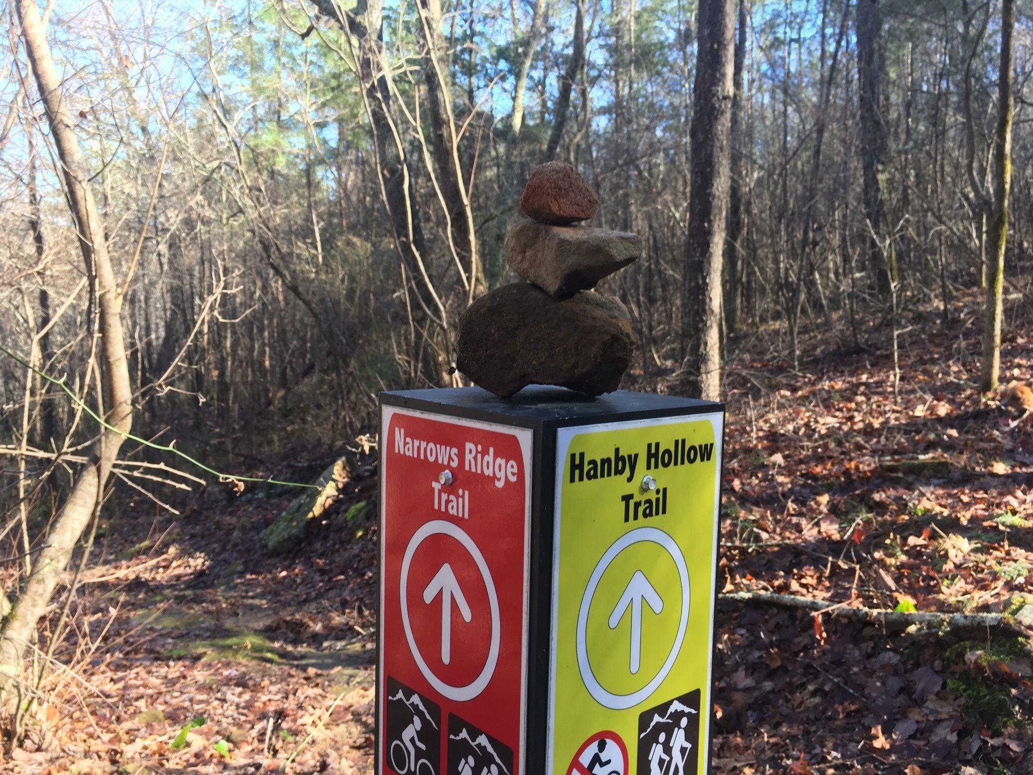 A trail marker showing directions for the Narrows Ridge Trail (red sign) and Hanby Hollow Trail (yellow sign), with a small stack of rocks balanced on top. The surrounding area is a wooded landscape with bare trees and fallen leaves. Narrow Ridge Loop Trail mountain bike trail.