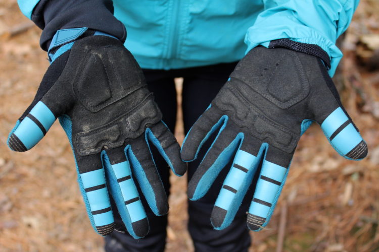 A pair of black and blue gloves held up by a person, displaying the palm and fingers. The gloves have a textured design with reinforced fingertips and a snug fit, suitable for outdoor activities. The background features a natural setting with pine needles on the ground.