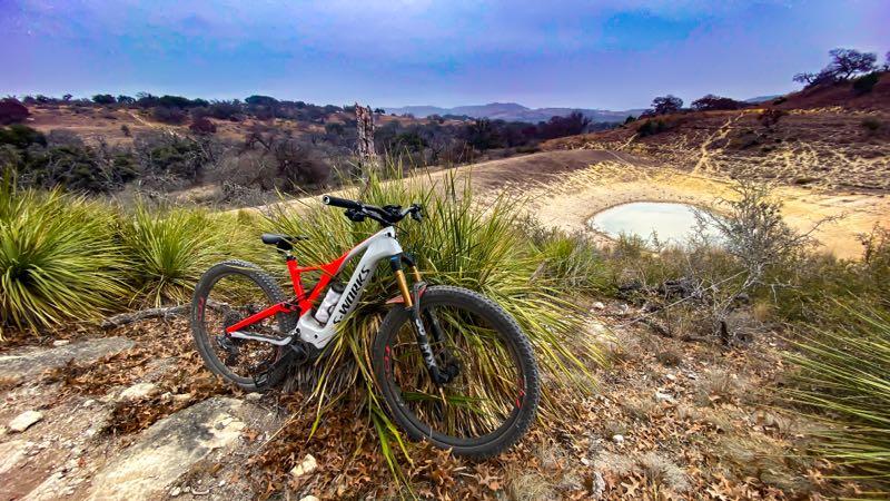 A mountain bike rests against a patch of tall grasses and rocky terrain, overlooking a dry landscape with a small pond in the distance. The scene is set under a cloudy sky, showcasing rolling hills and sparse vegetation typical of a natural outdoor environment. Flat Rock Ranch mountain bike trail.