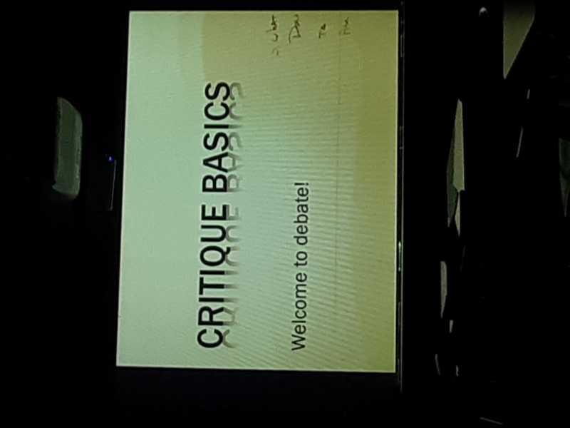 A presentation slide titled "CRITIQUE BASICS" with the subtitle "Welcome to debate!" displayed on a screen. The slide features a light background with bold black text. Big Creek mountain bike trail.