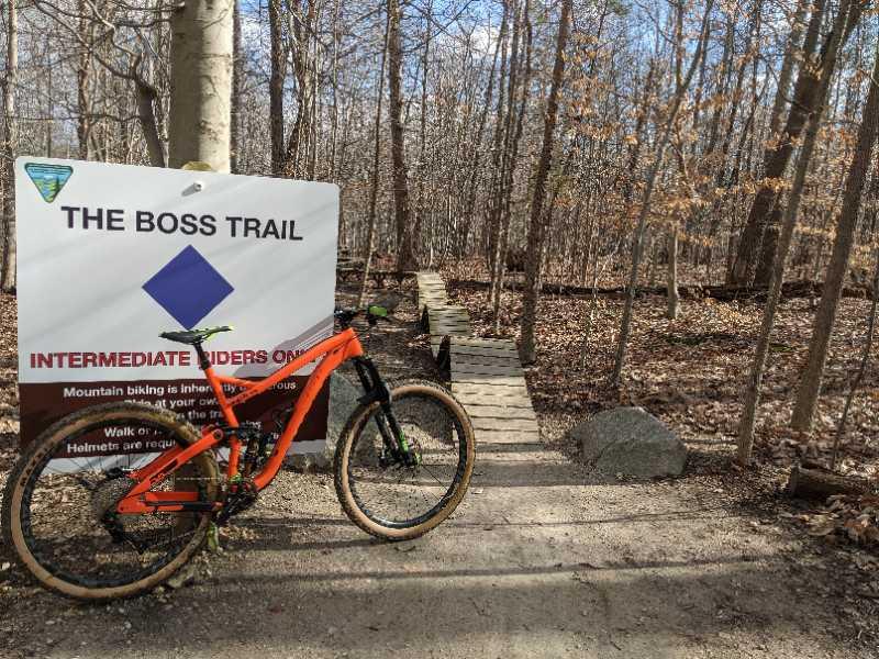 A mountain bike is leaning against a trail sign labeled "THE BOSS TRAIL," indicating that it is for intermediate riders only. The sign includes instructions about mountain biking safety. In the background, there are trees with sparse foliage, suggesting an early spring or late autumn setting. A wooden pathway leads into the forest, hinting at an adventurous cycling route ahead. Meadowood mountain bike trail.