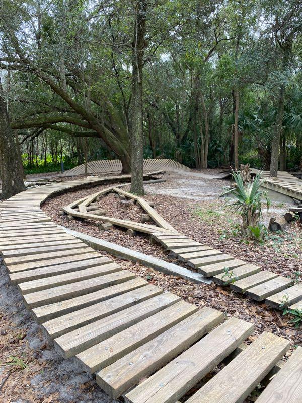 A winding wooden boardwalk path meanders through a lush green forest, surrounded by trees and foliage. The path features various curves and shapes, with some sections elevated or lined with leaves, creating a natural and tranquil atmosphere. Mount Dora Trail mountain bike trail.