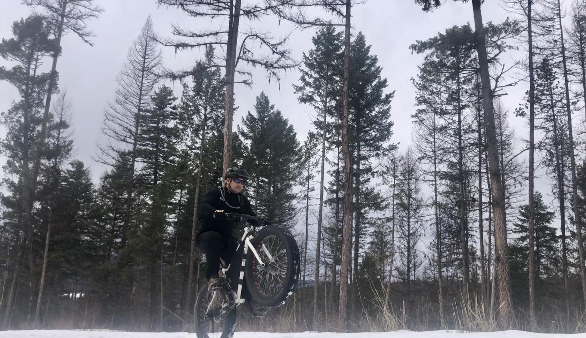 A mountain biker performing a wheelie on a snowy terrain, surrounded by tall coniferous trees under a cloudy sky. Spencer Mountain mountain bike trail.